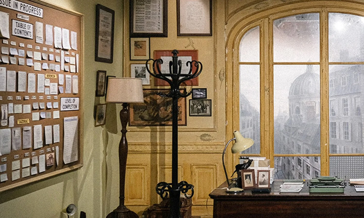 An office with a bulletin board, various framed pictures, a floor lamp, a coat rack, a desk with a lamp and papers, and large windows overlooking historic buildings.