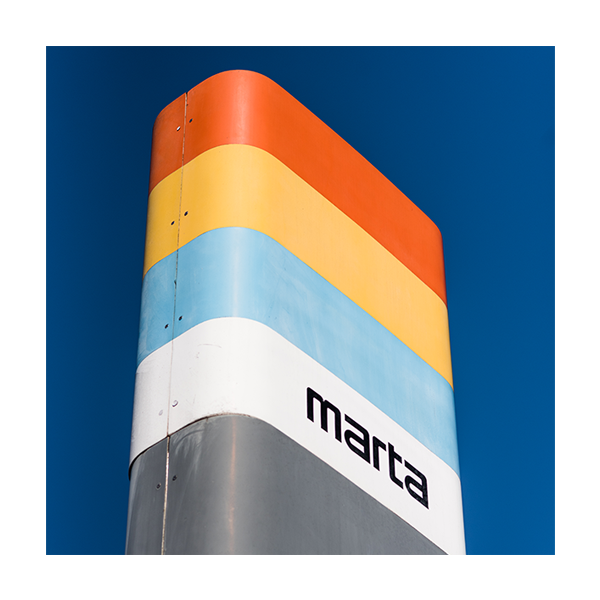 Close-up of a colorful sign with red, yellow, blue, white, and gray stripes, displaying the text "marta" against a clear blue sky.