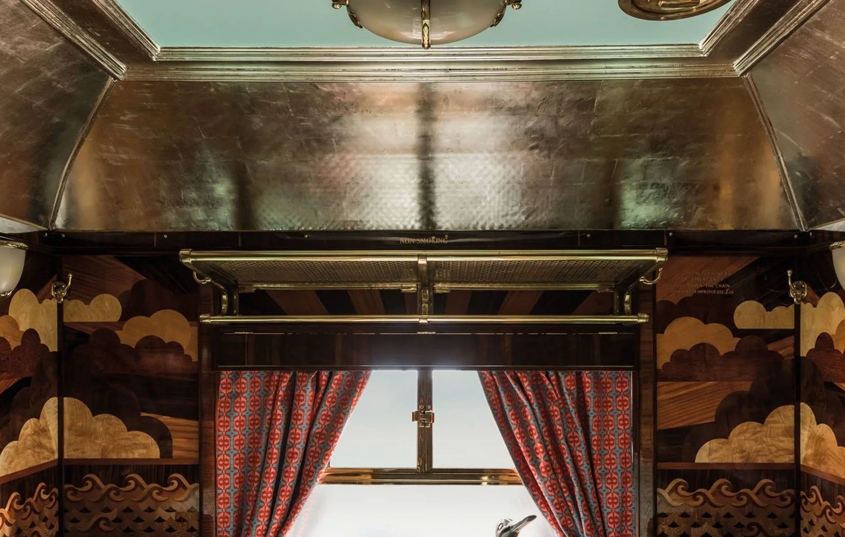 Interior of a vintage train car with patterned wooden walls, red patterned curtains, a window, and a metallic overhead luggage rack.