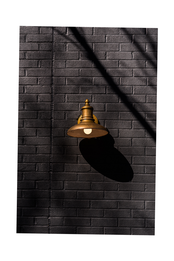 A vintage-style brass wall-mounted outdoor light fixture casting a shadow on a dark gray brick wall.