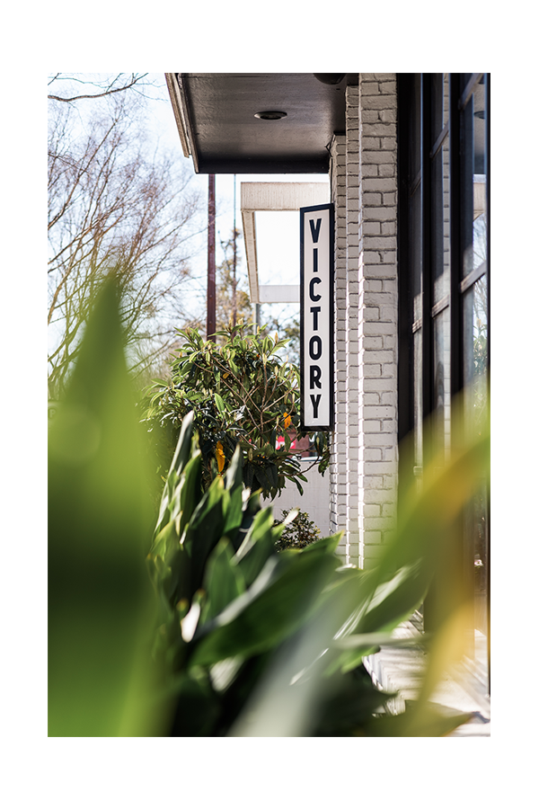 A vertical sign with the word "VICOTRY" on a white brick building, partially obscured by green plants and trees outside on a clear day.
