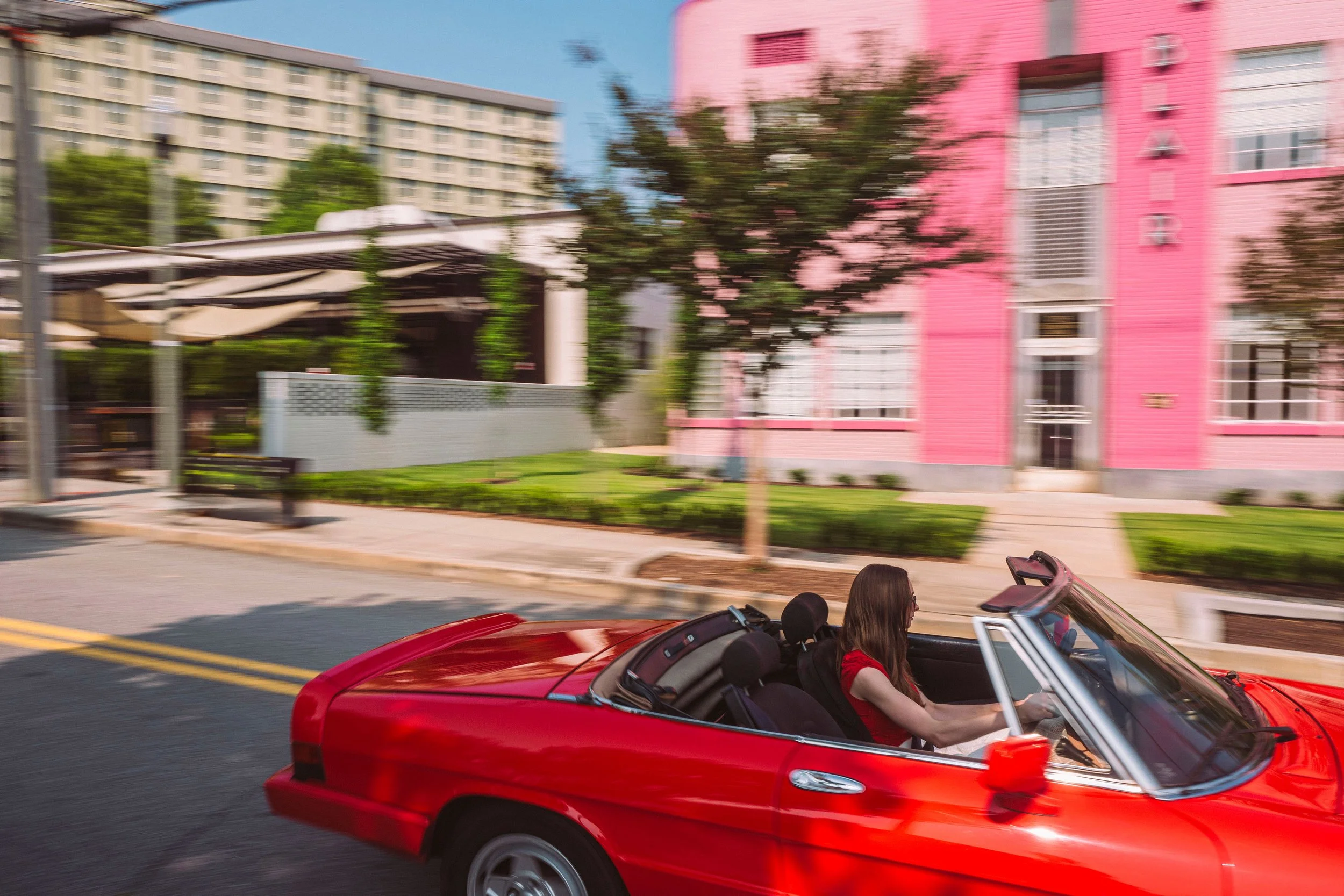 A woman with long brown hair wearing a red shirt driving a red convertible car on a city street with colorful buildings in the background, motion-blurred to show movement.