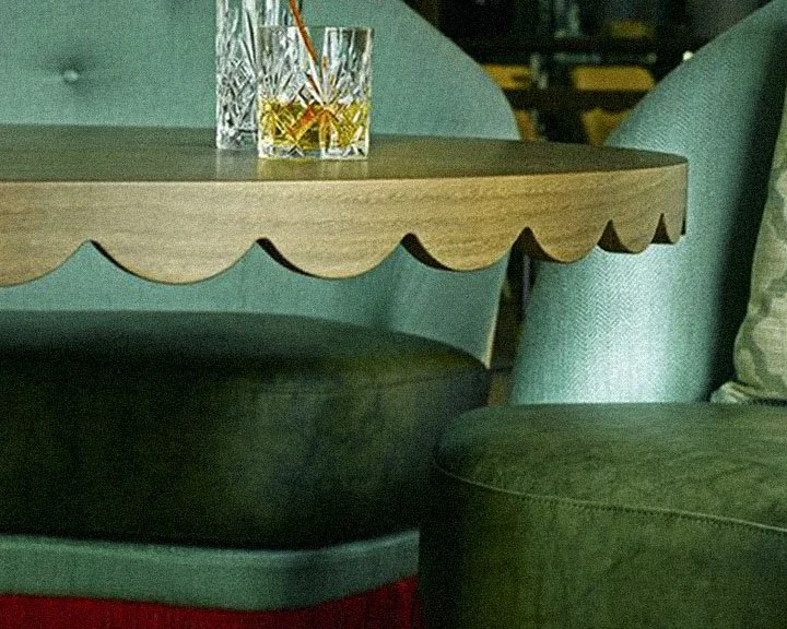 Two glasses of whiskey on a wooden table near a teal-colored upholstered chair in a cozy indoor setting.