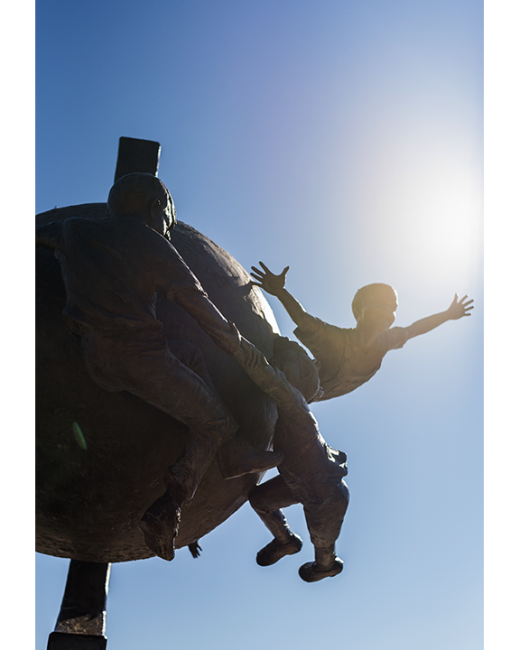 A sculpture of a person being helped up a rock wall, with one arm outstretched, against a bright blue sky and sunlight.