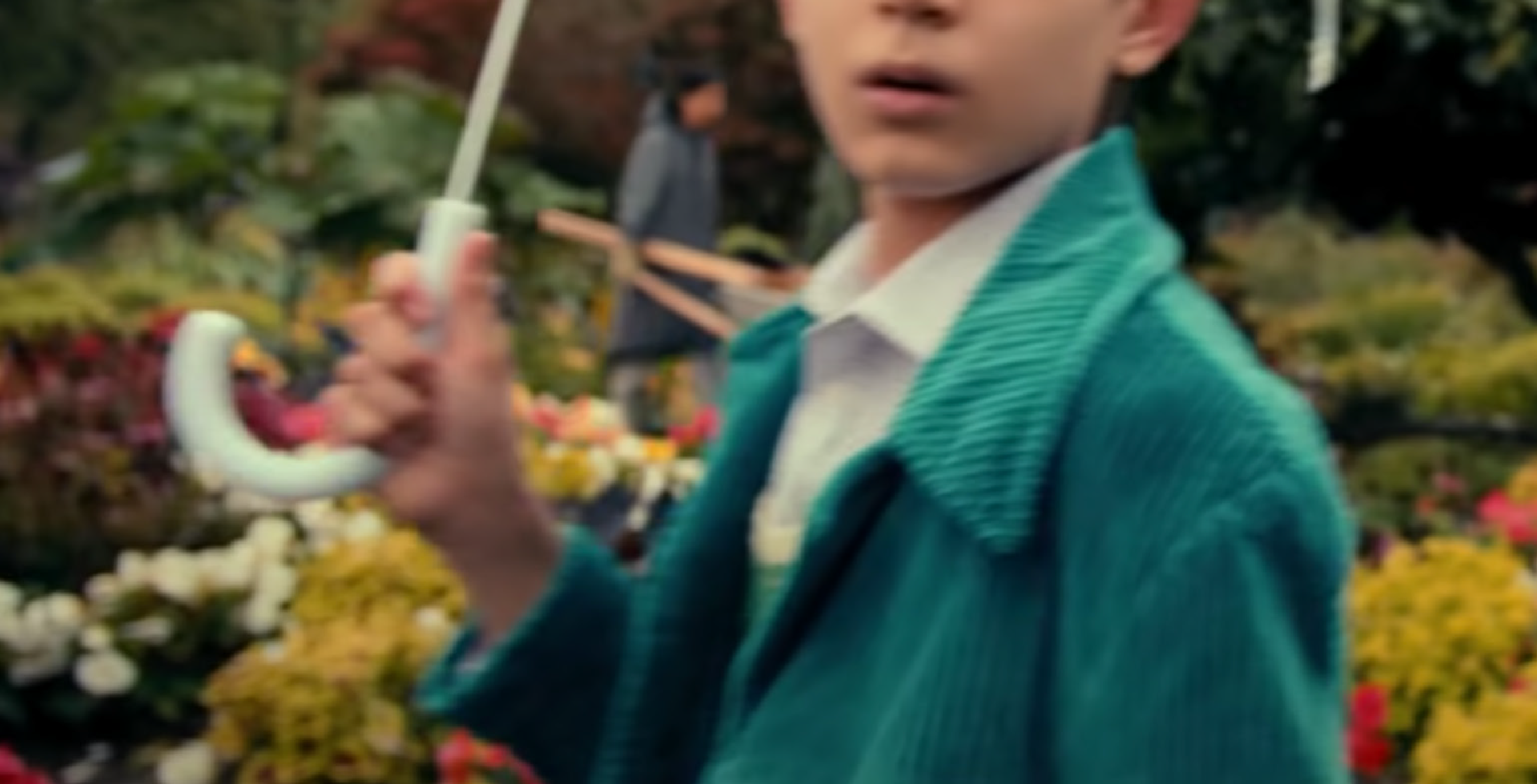 A young boy in a teal sweater holding a white umbrella-shaped object with a pink handle, standing in a garden or park with flowers and trees in the background.