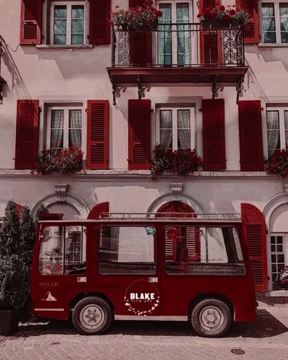 Red vintage van parked in front of a European-style building with red shutters and flower boxes.