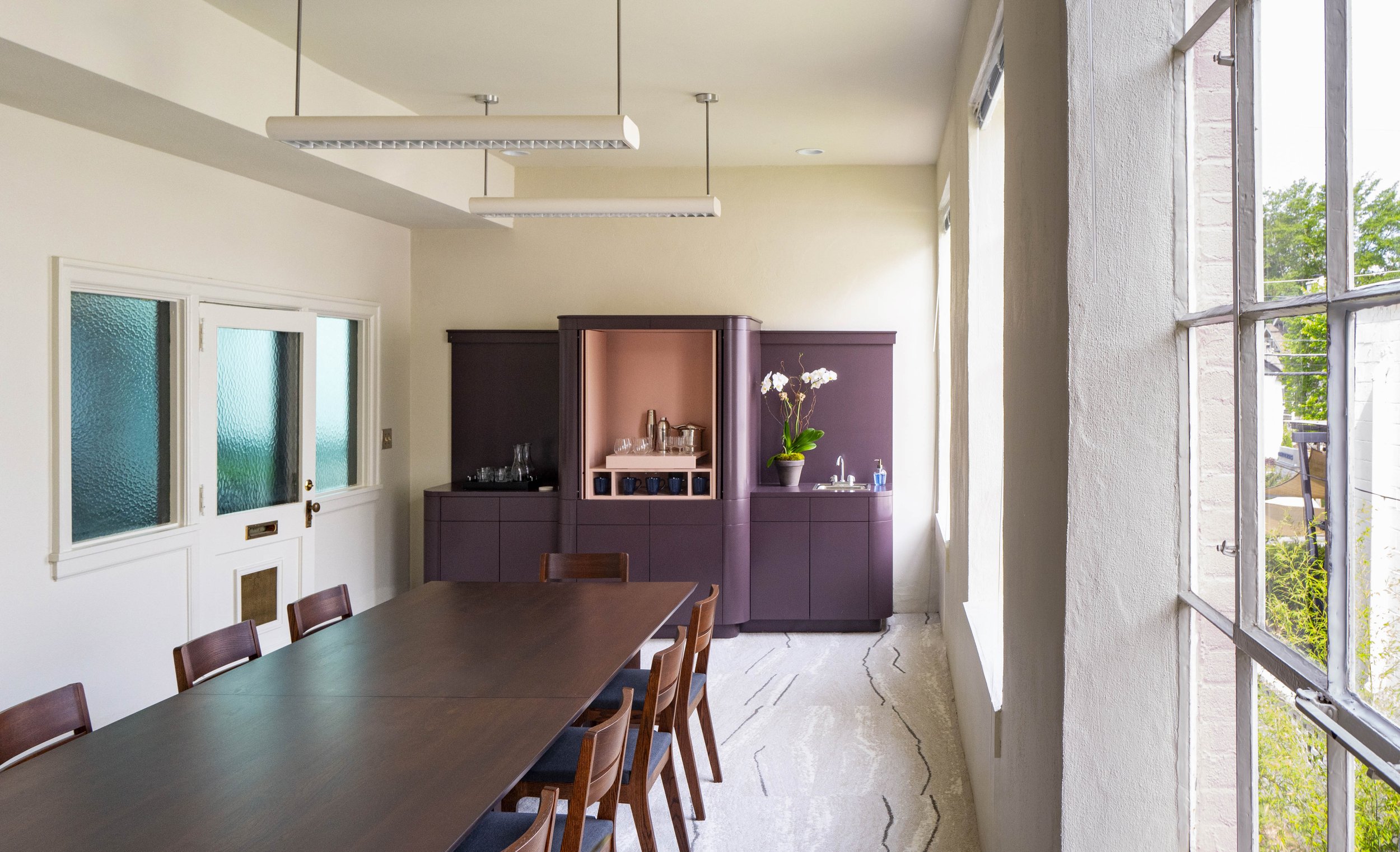 Bright dining area with large window, a long wooden table, and purple cabinetry with glassware and a potted orchid.