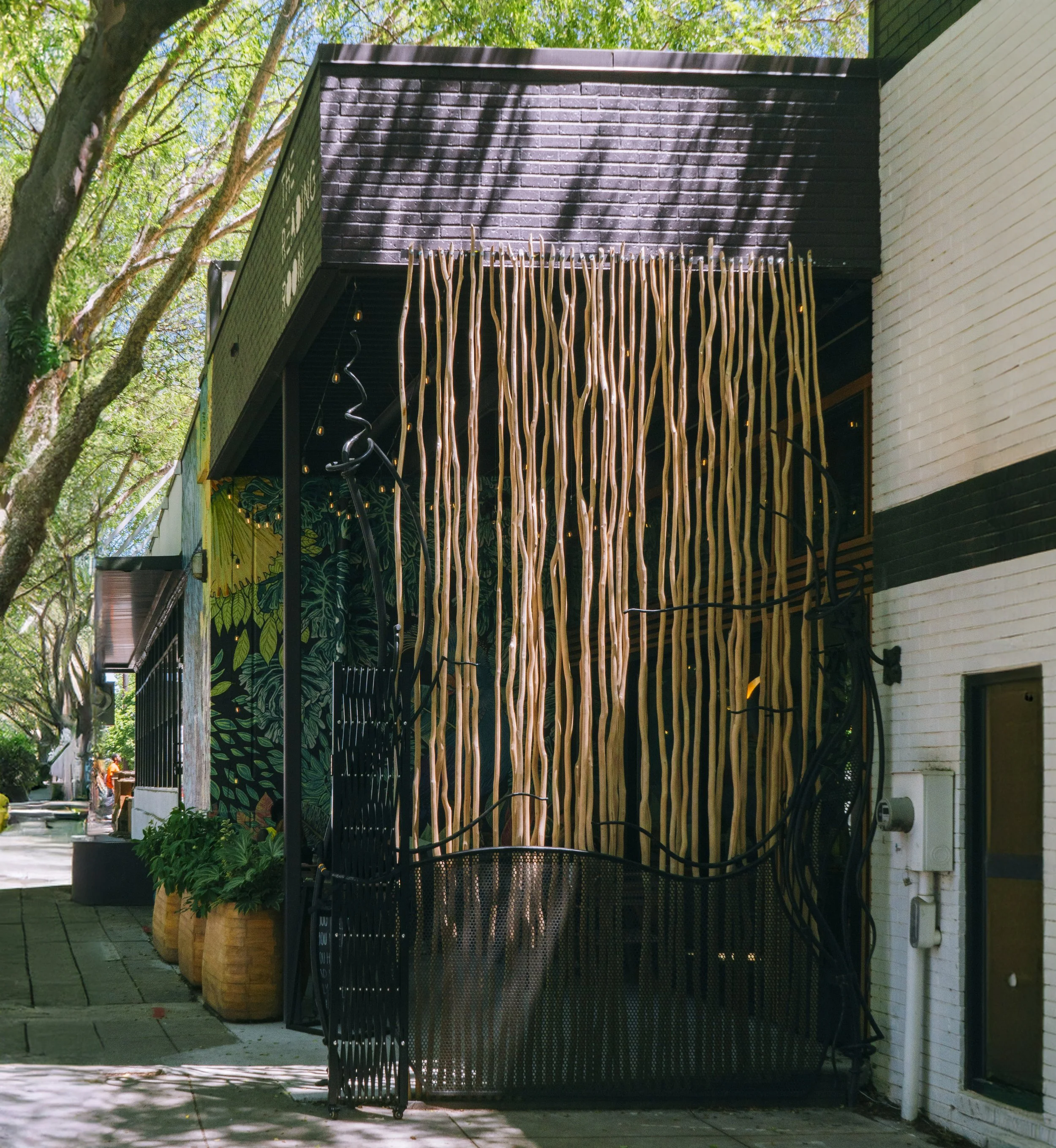 An artistic installation on the exterior of a building features wooden vertical slats, black metal branches, and green foliage designs on the wall. The building is white brick with a black trim, with large potted plants nearby and trees providing sha