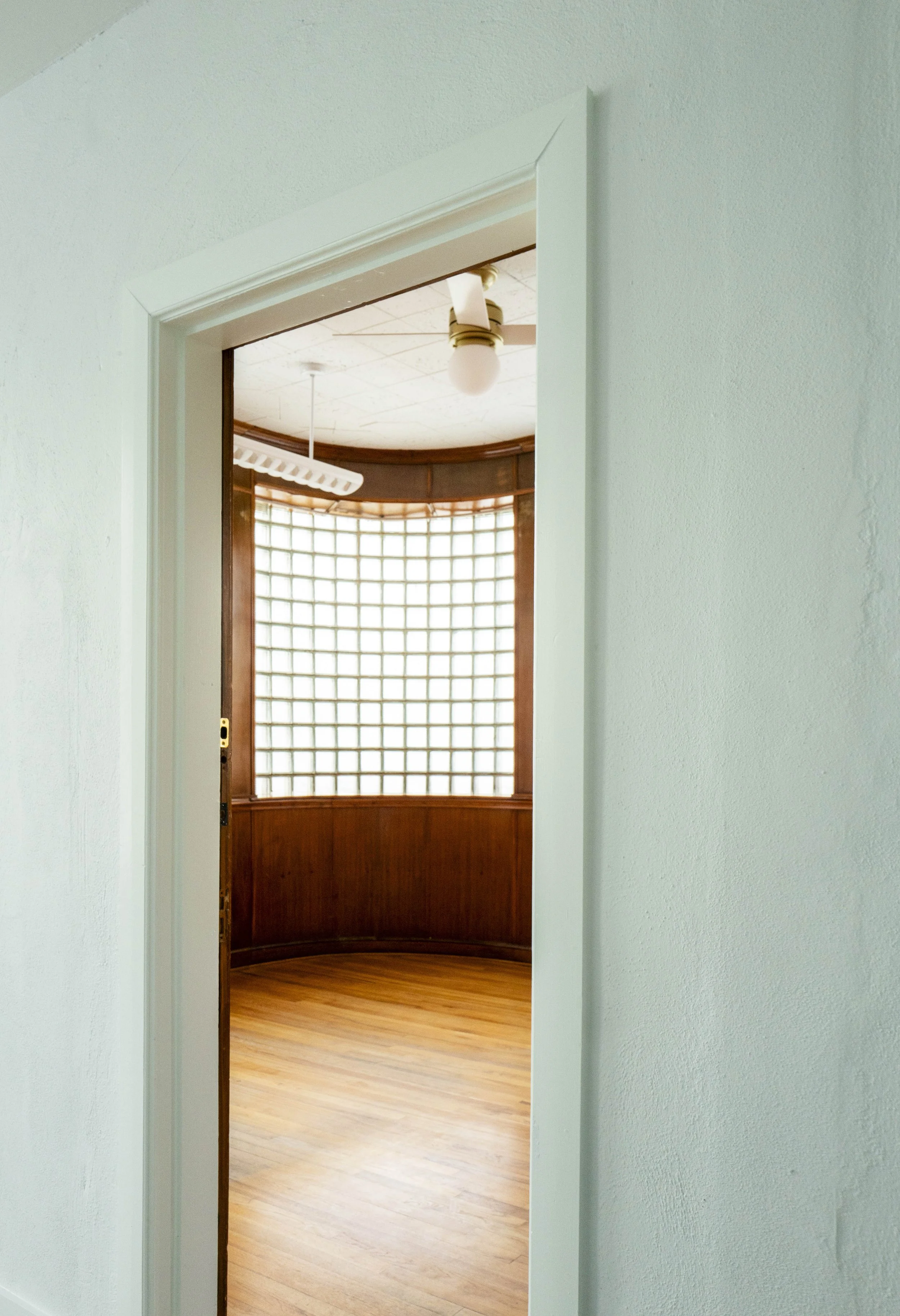 Interior view of a room with a circular glass block wall, wooden paneling, a hardwood floor, and a ceiling with a light fixture and a hanging drying rack.