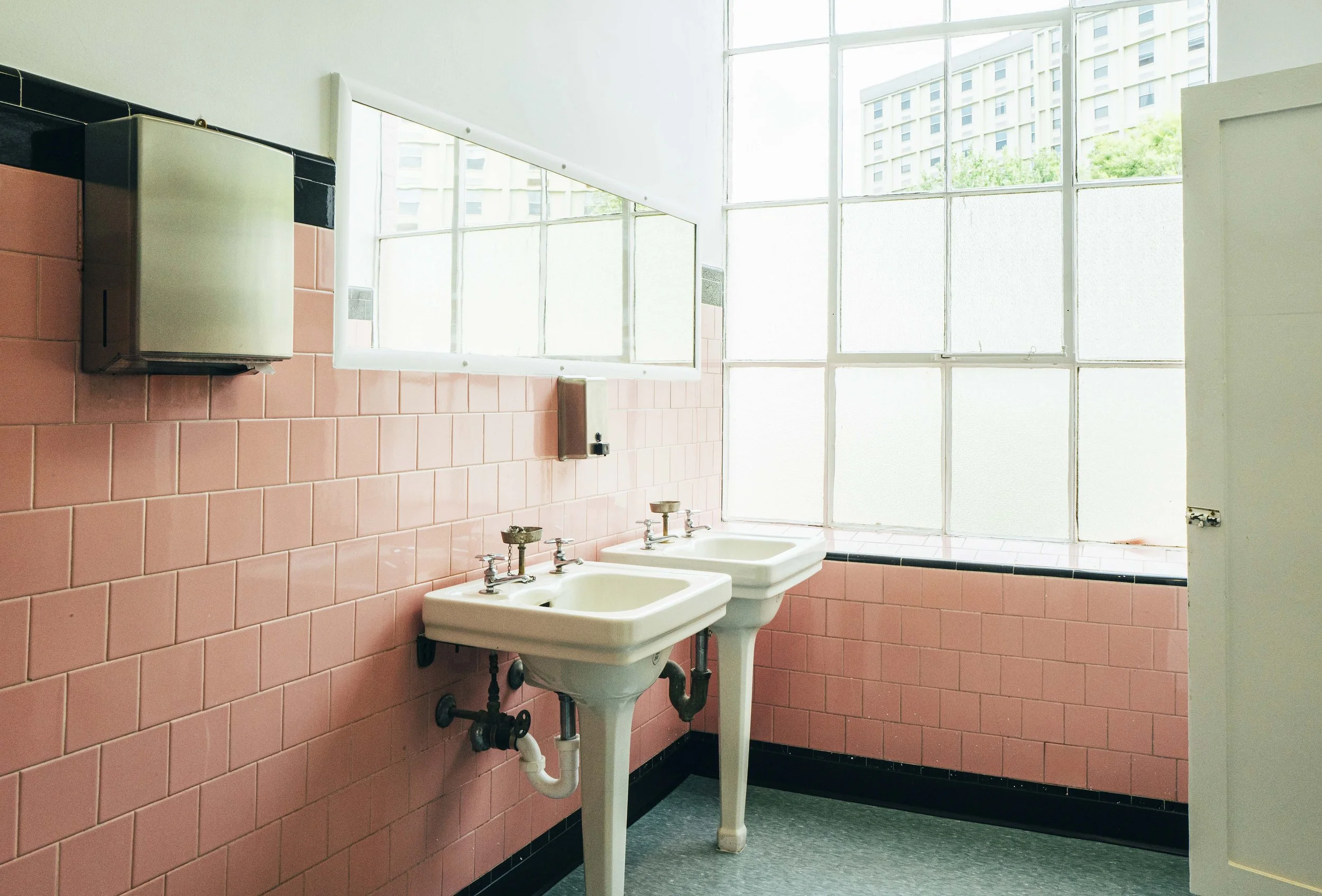 Public restroom with two white sinks, pink tiled wall, and large frosted window providing natural light.