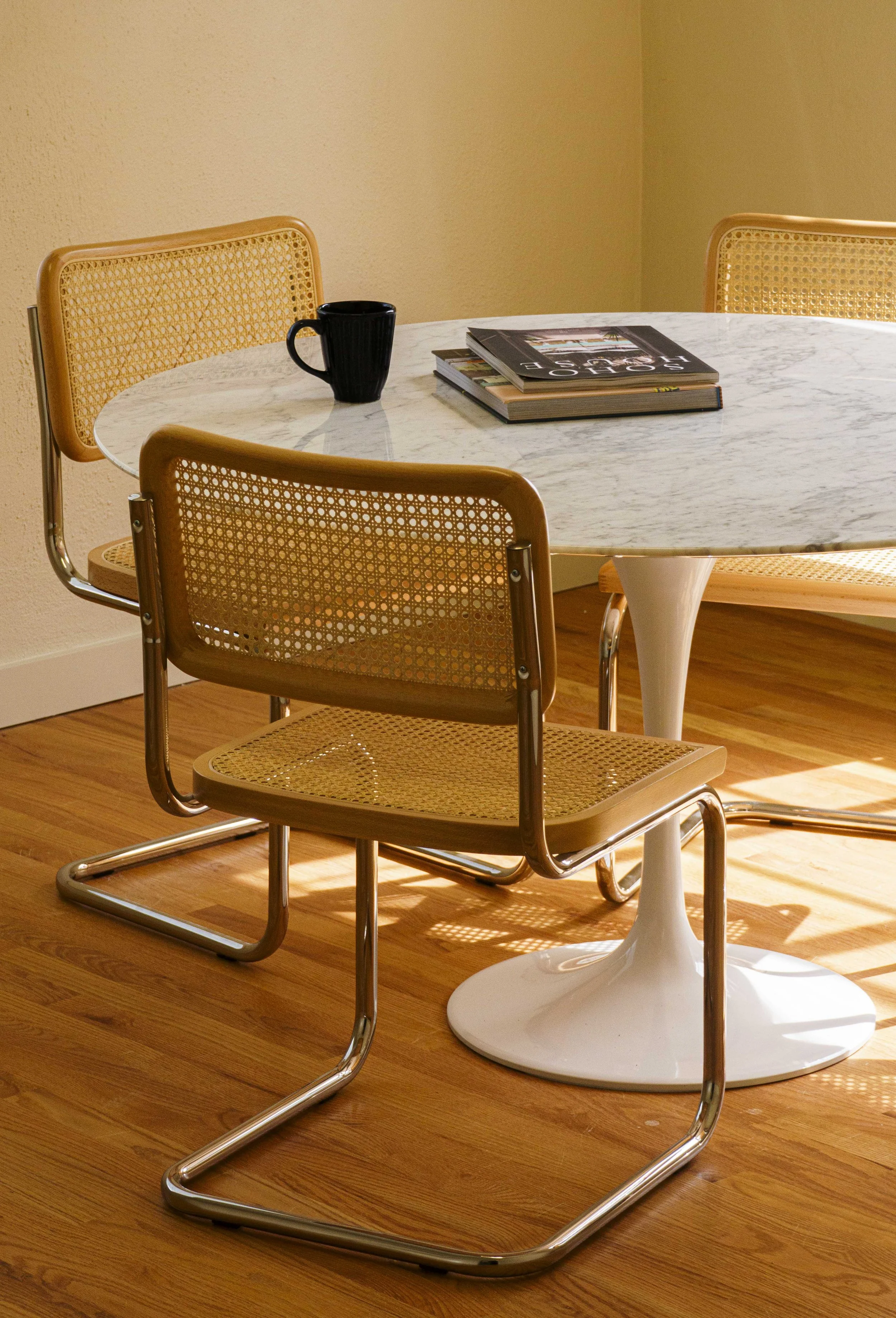 A round marble dining table with three wooden and metal chairs, a black mug, and a stack of magazines on top, placed on a wooden floor with a beige wall in the background.