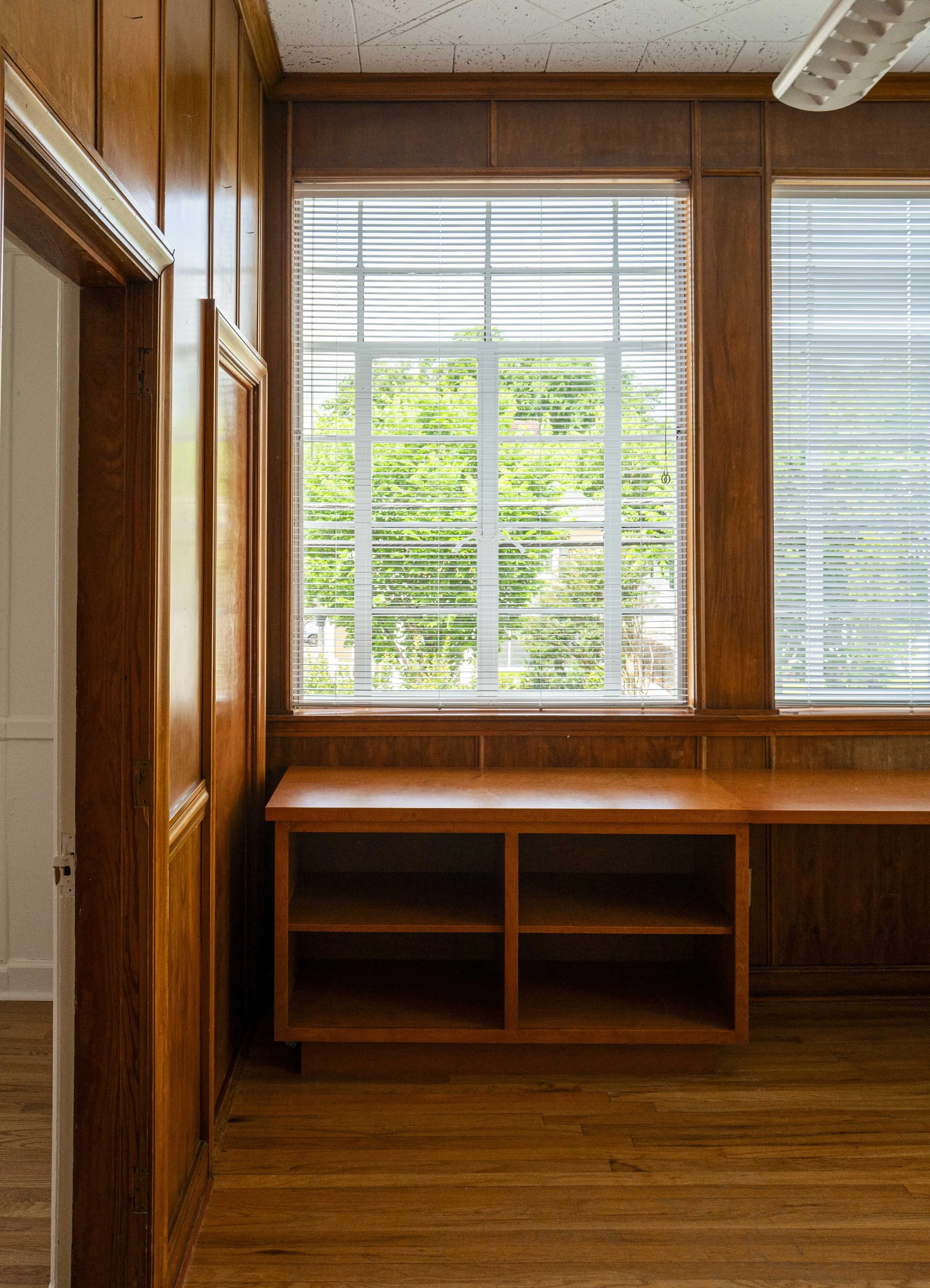 A wood-paneled room with a large window showing greenery outside. There is a built-in wooden cabinet with open shelves beneath the window, and horizontal blinds covering the window.
