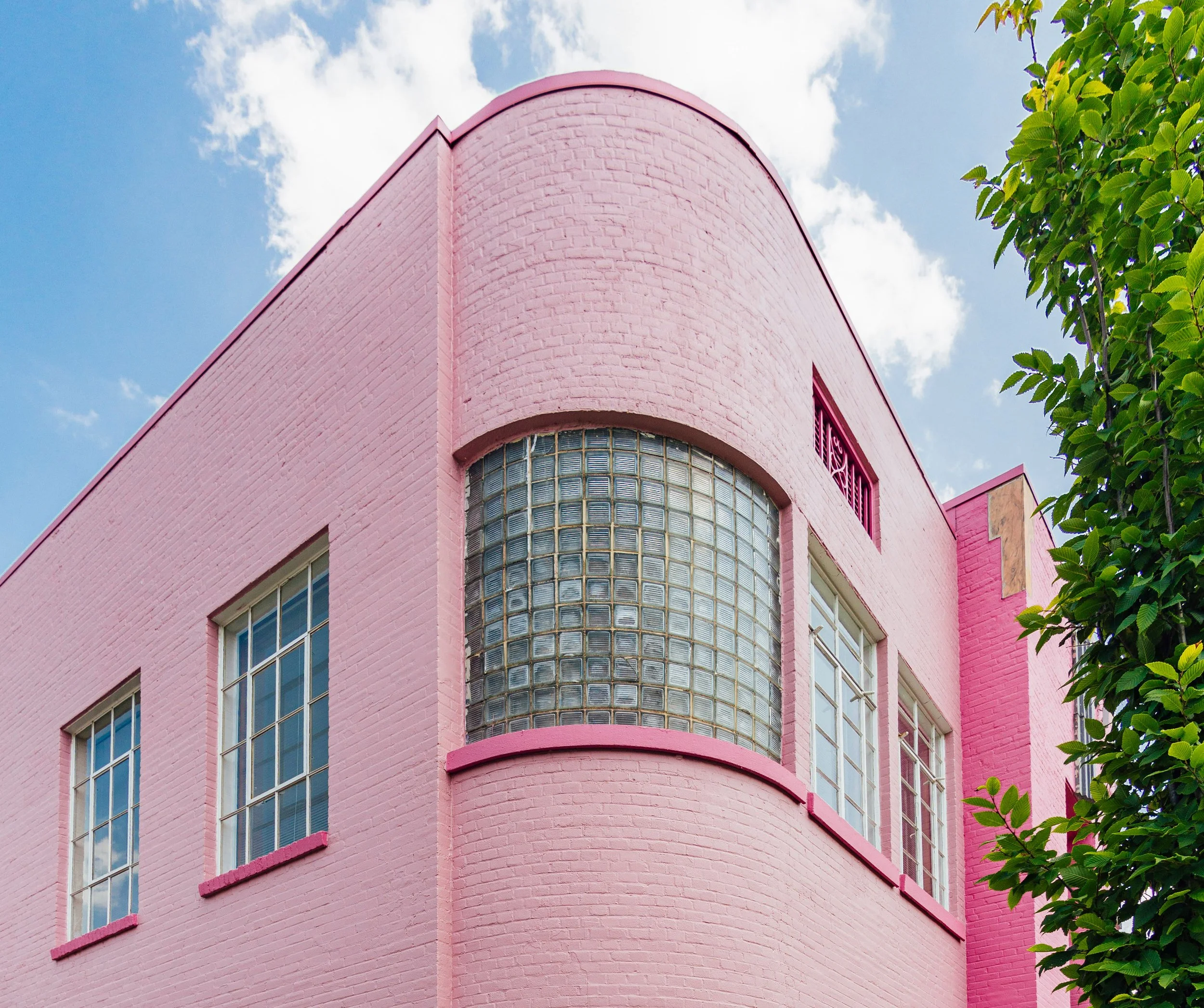 A pink building with large glass block windows and multiple rectangular windows, with a clear blue sky and some clouds in the background. A leafy green tree is on the right side of the image.