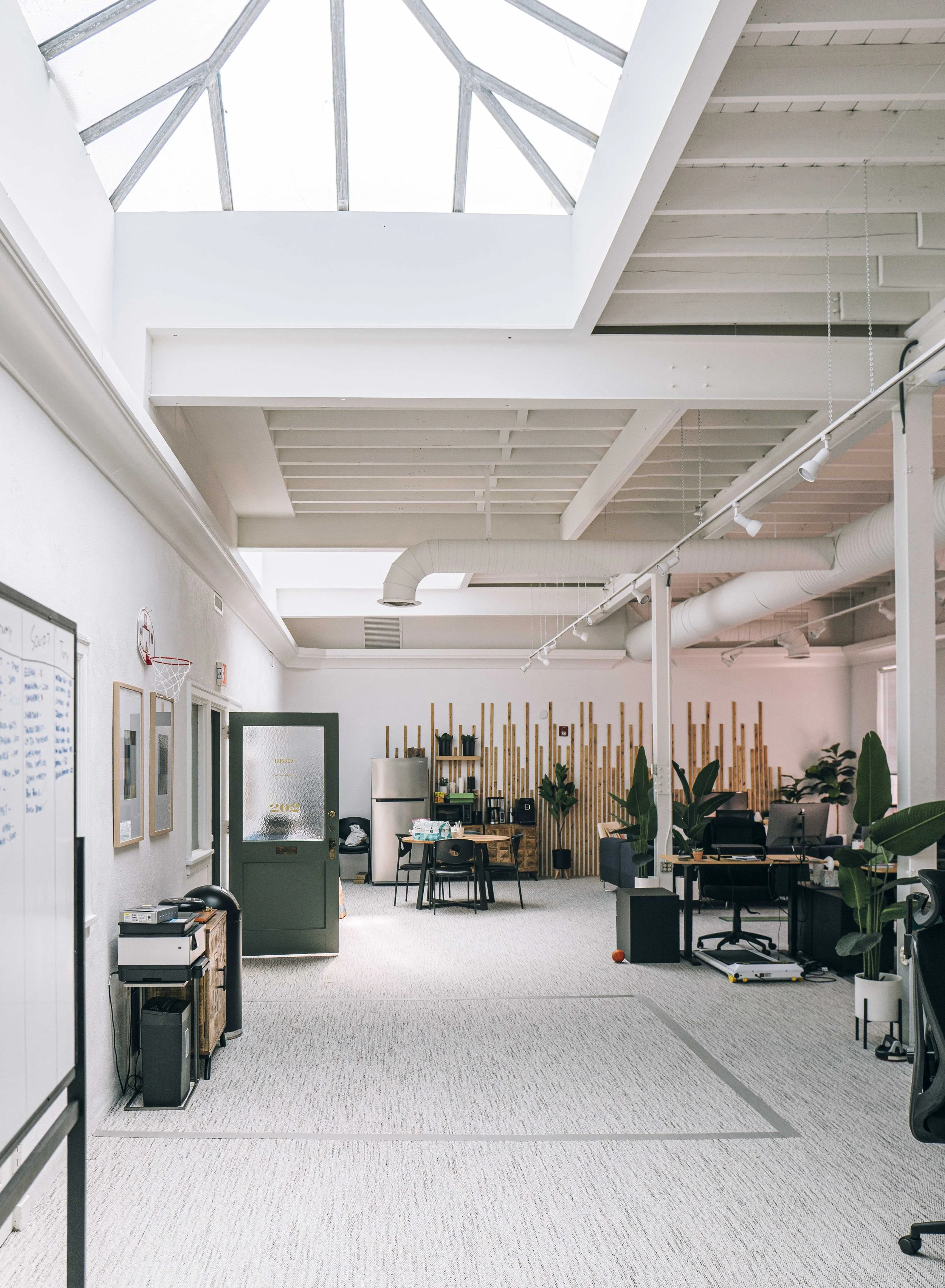 Open-plan office space with workstations, plants, a mini refrigerator, and natural light from a large skylight overhead.
