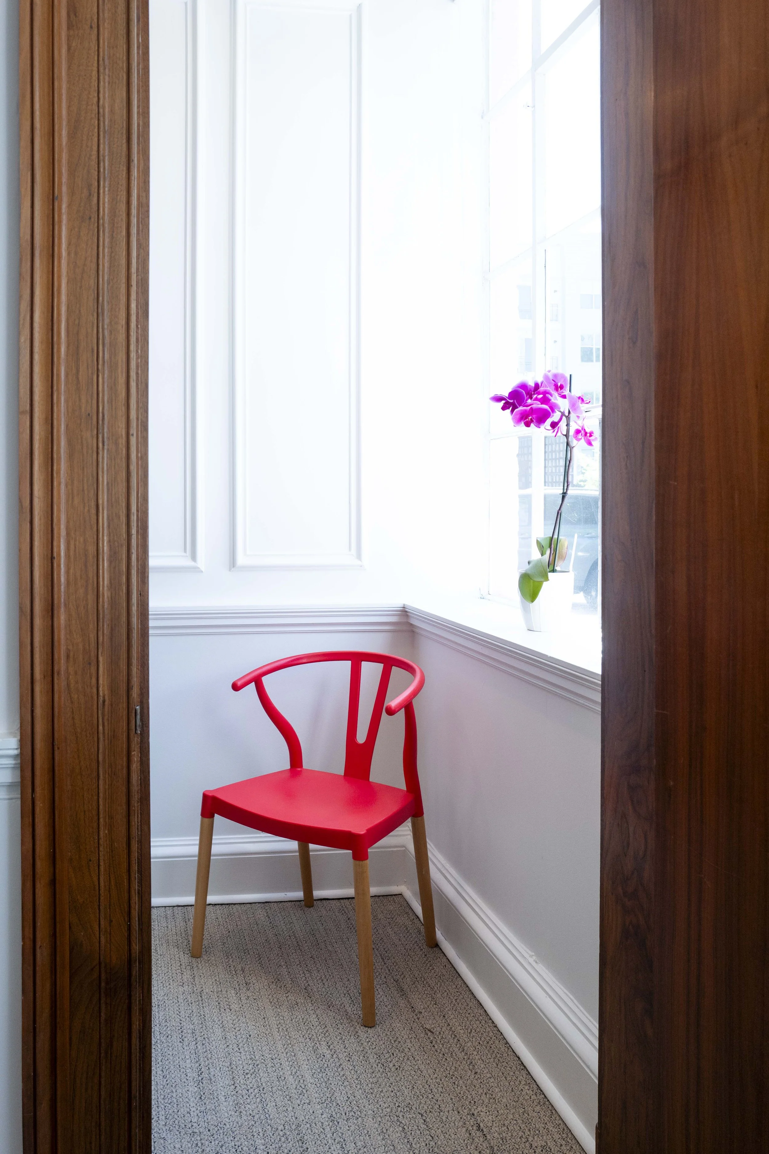 A small red chair with wooden legs placed in a corner near a large window, with a pink orchid in a white pot on the windowsill and white walls adorned with decorative trim.