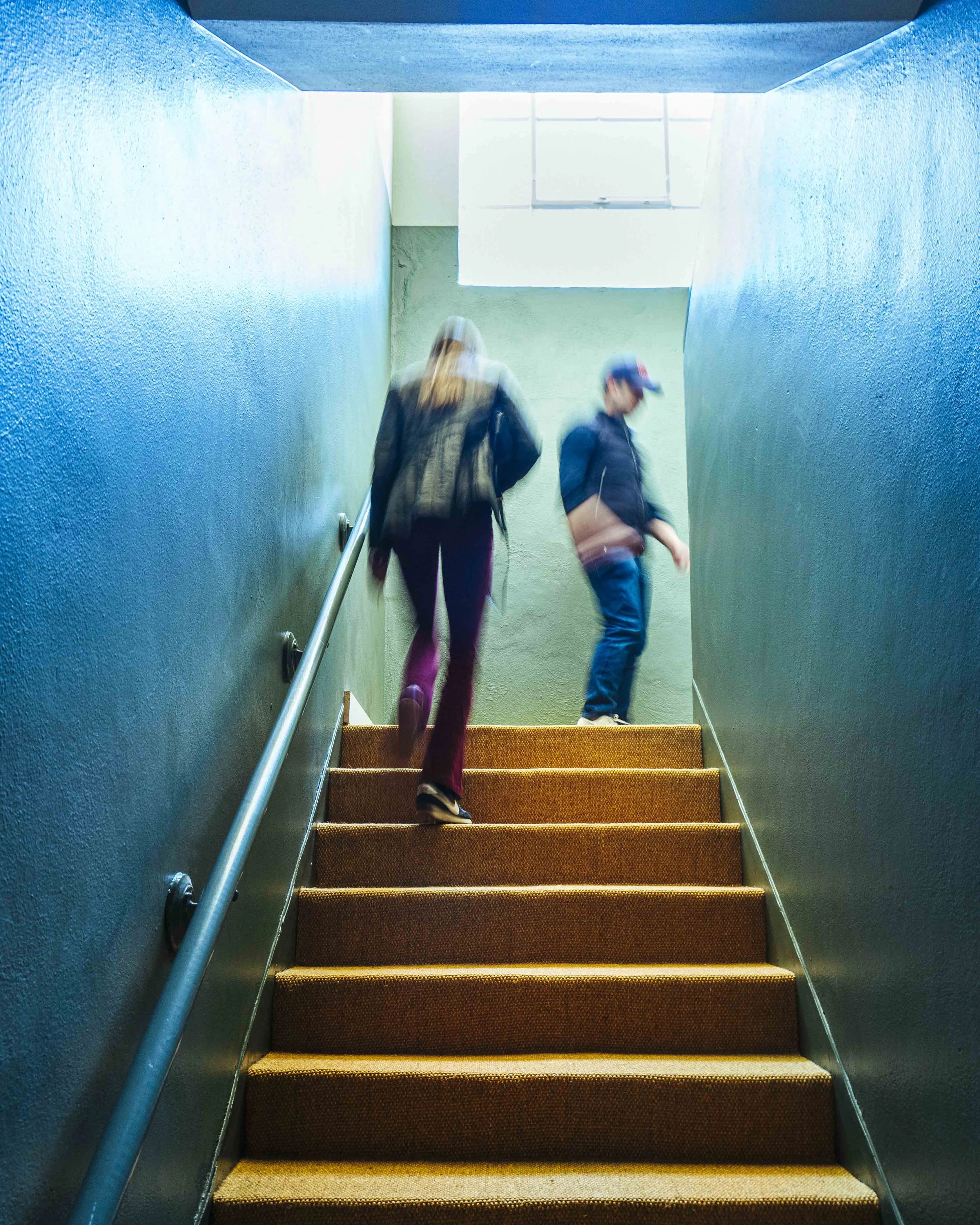 Two people walking up a staircase with blue green walls and orange carpeted steps.