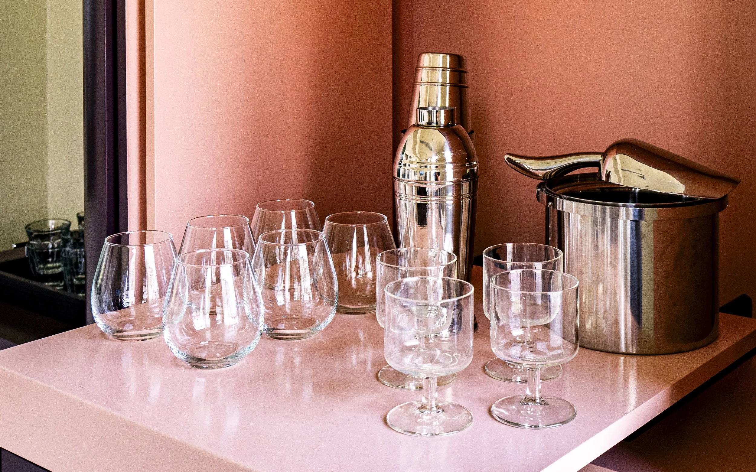 A pink table with a stainless steel cocktail shaker, a chiller bucket, a beverage dispenser, and nine empty glassware including cocktail glasses and wine glasses.