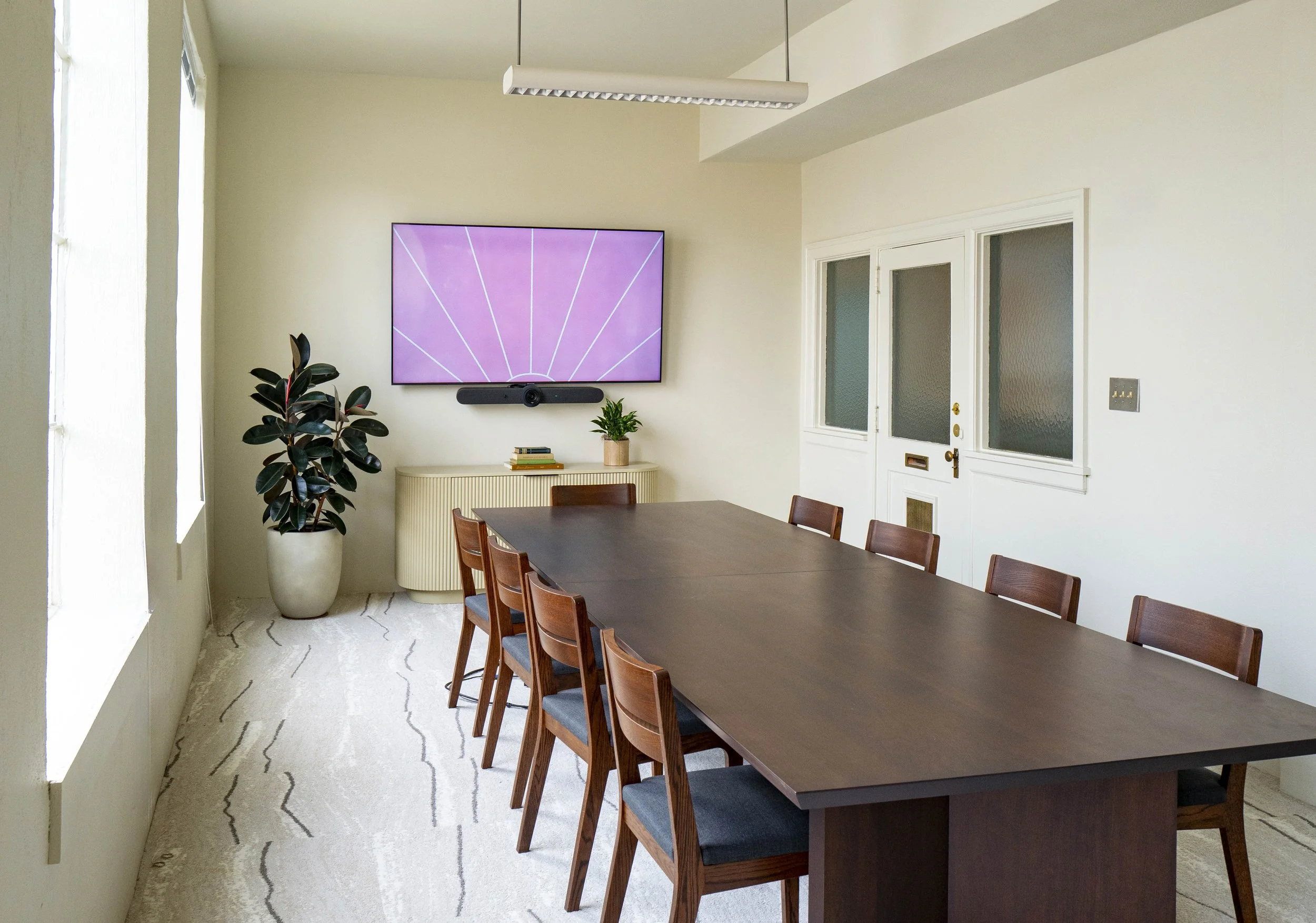 Empty conference room with large wooden table, eight chairs, a wall-mounted monitor, speaker, and potted plants.