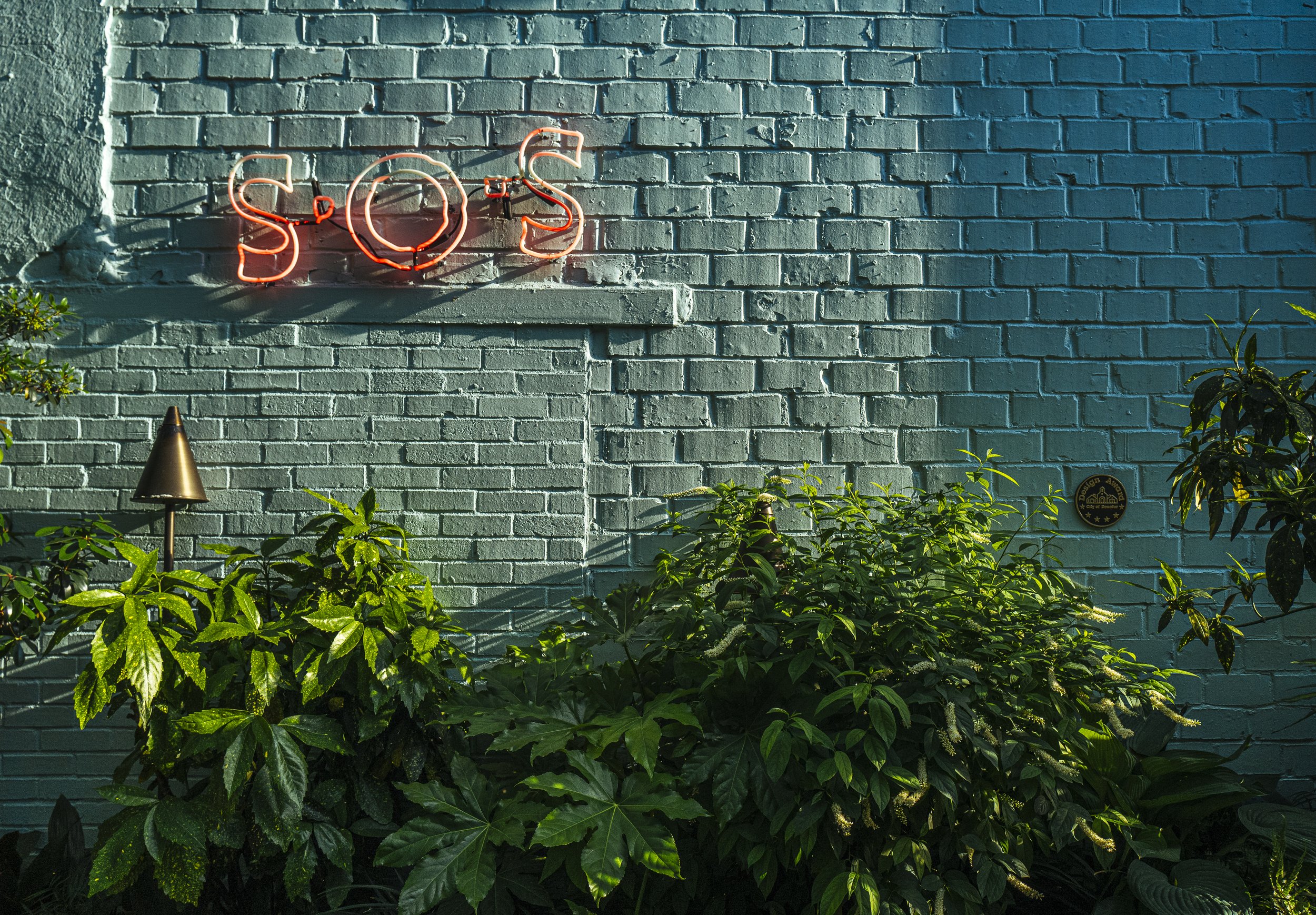 Neon sign spelling 'S.O.S' mounted on a teal brick wall, with lush green foliage and a small garden light below.