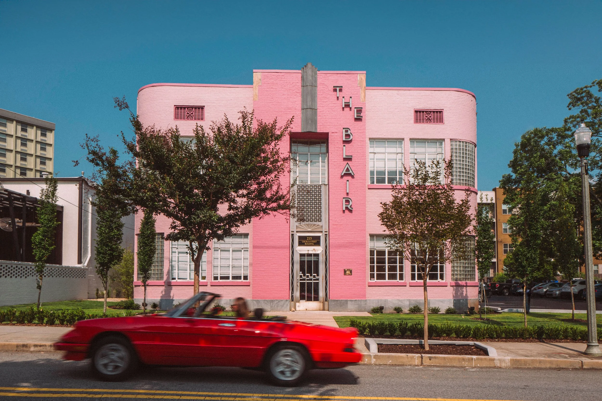 A pink art deco building with the word "THE BLAIR" vertically on the front, trees lining the sidewalk, and a red convertible car driving by in the foreground.