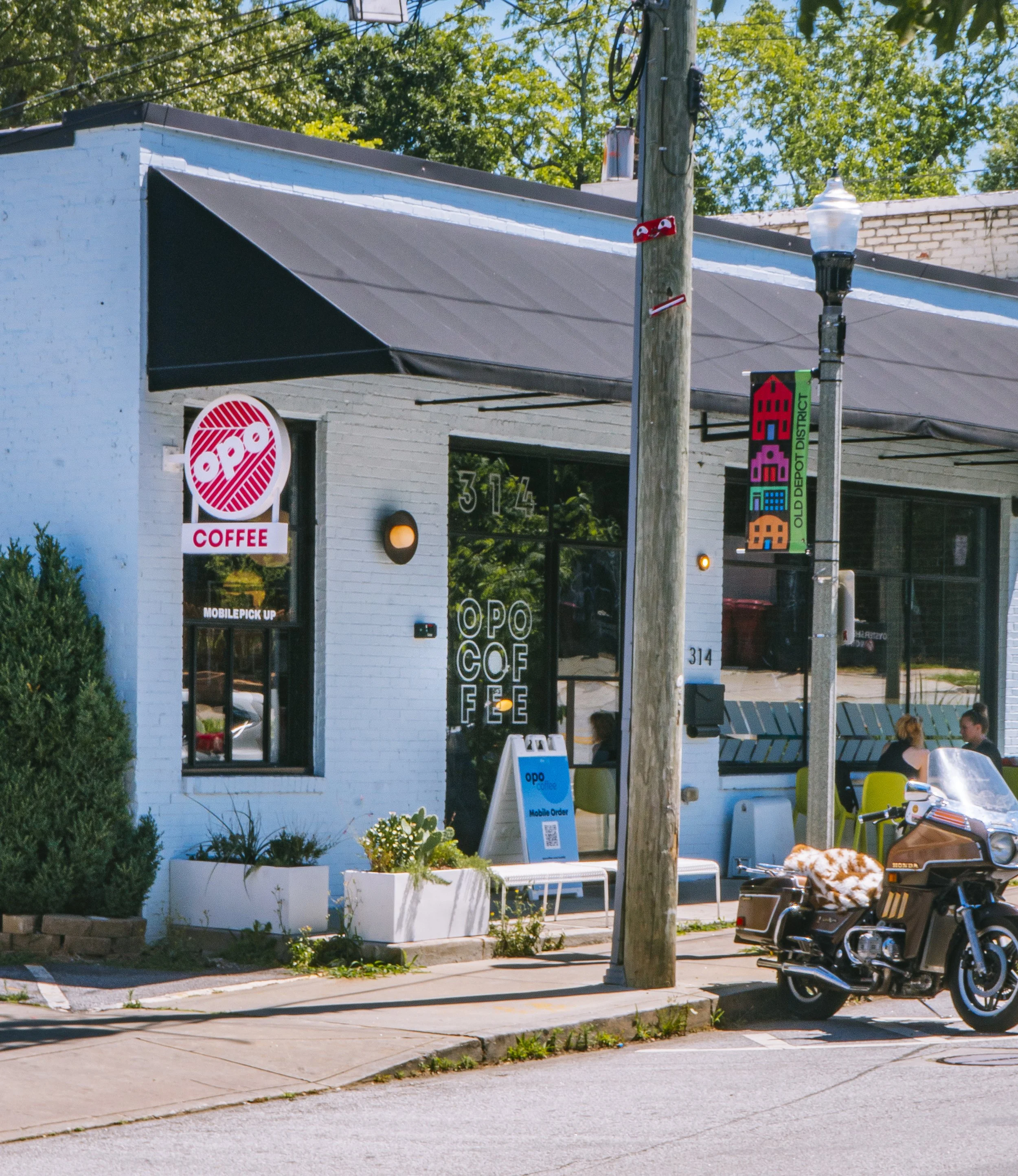Exterior of a coffee shop named OPO Coffee with large glass windows, a sign with the name, and outdoor seating. A motorcycle is parked in front, and there are trees in the background.
