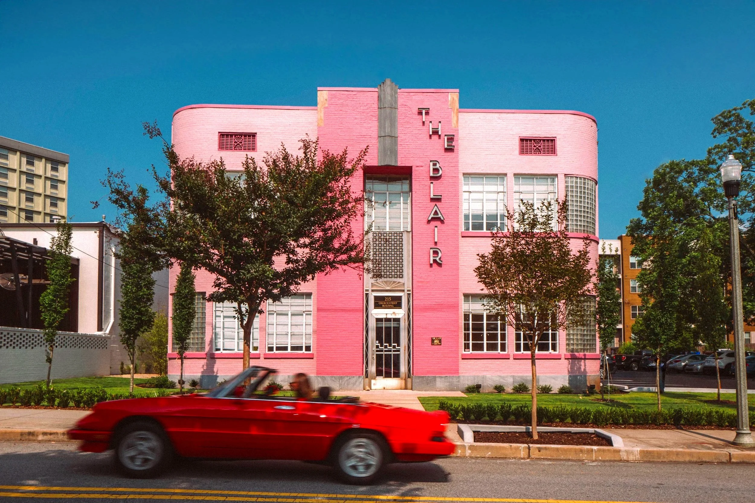 Pink Art Deco building labeled The Blair with a red convertible driving by in the foreground under a clear blue sky, with trees and street lamps lining the sidewalk.