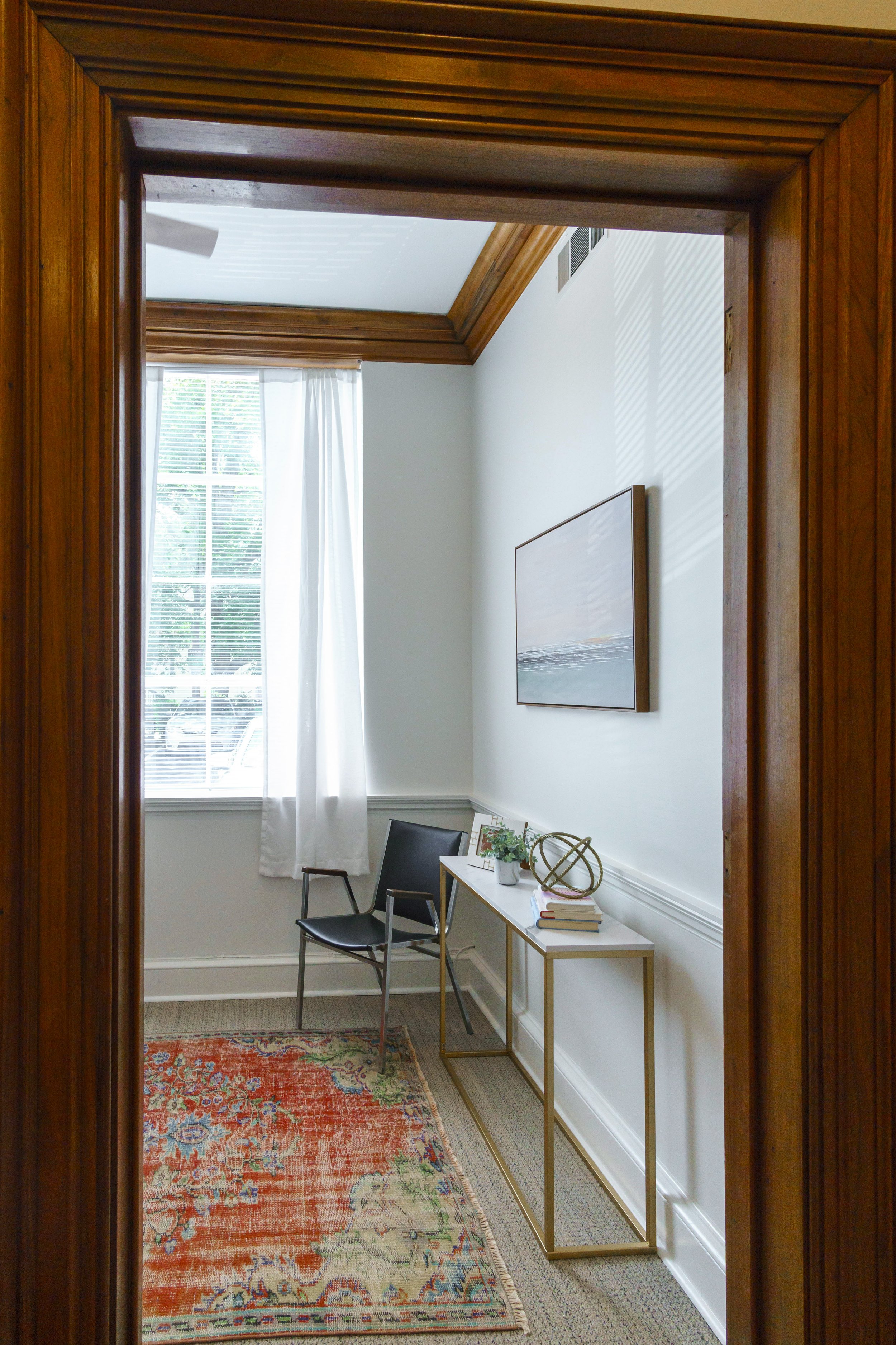 A small interior room with a large window with white curtains, a black chair, a white table with gold legs, decorative items, and a colorful rug, viewed through a wooden doorway.