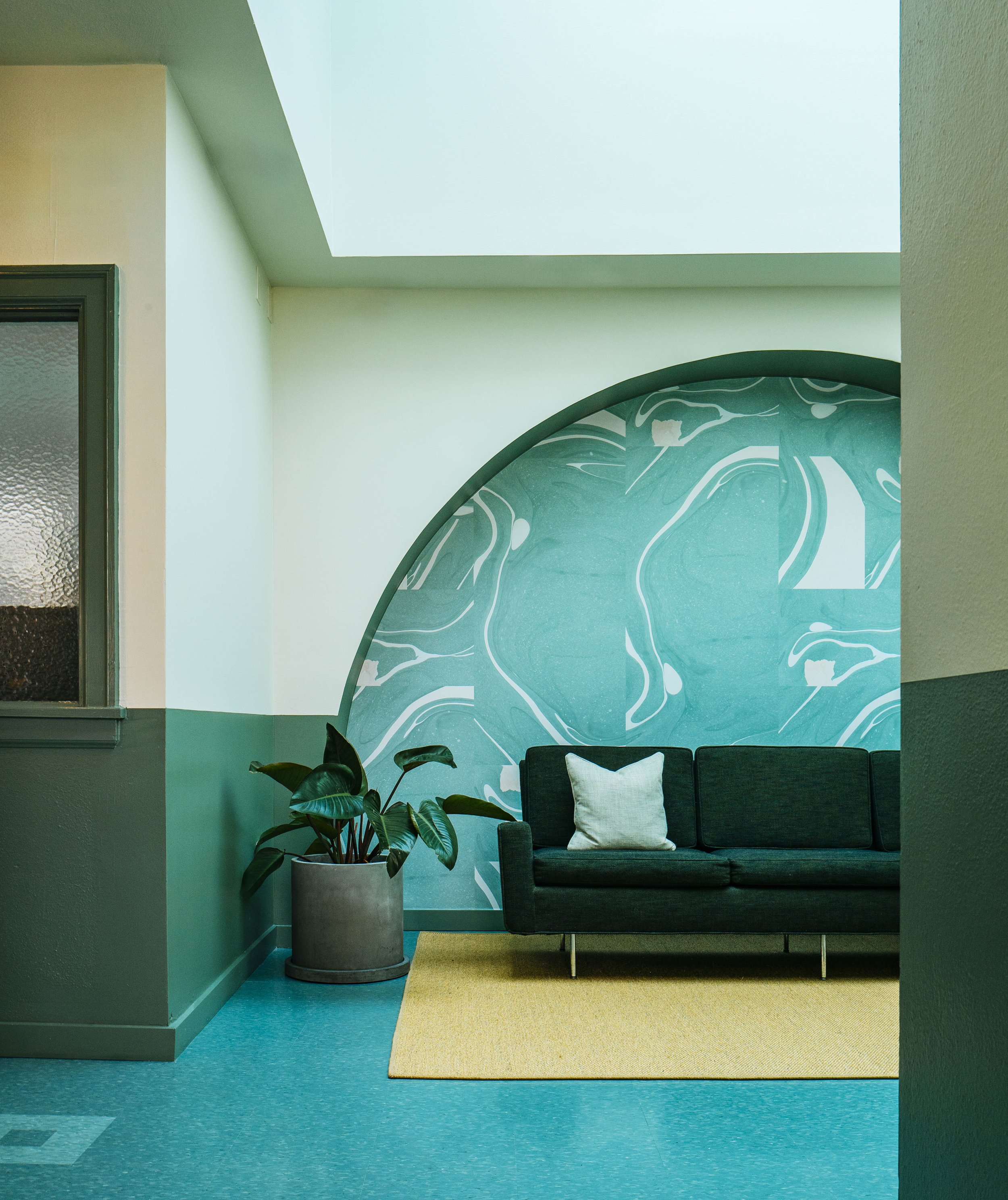A modern indoor space with a teal and white abstract wall mural, a black sofa with a white pillow, a potted plant, and a beige rug on teal flooring, illuminated by natural light from a skylight.