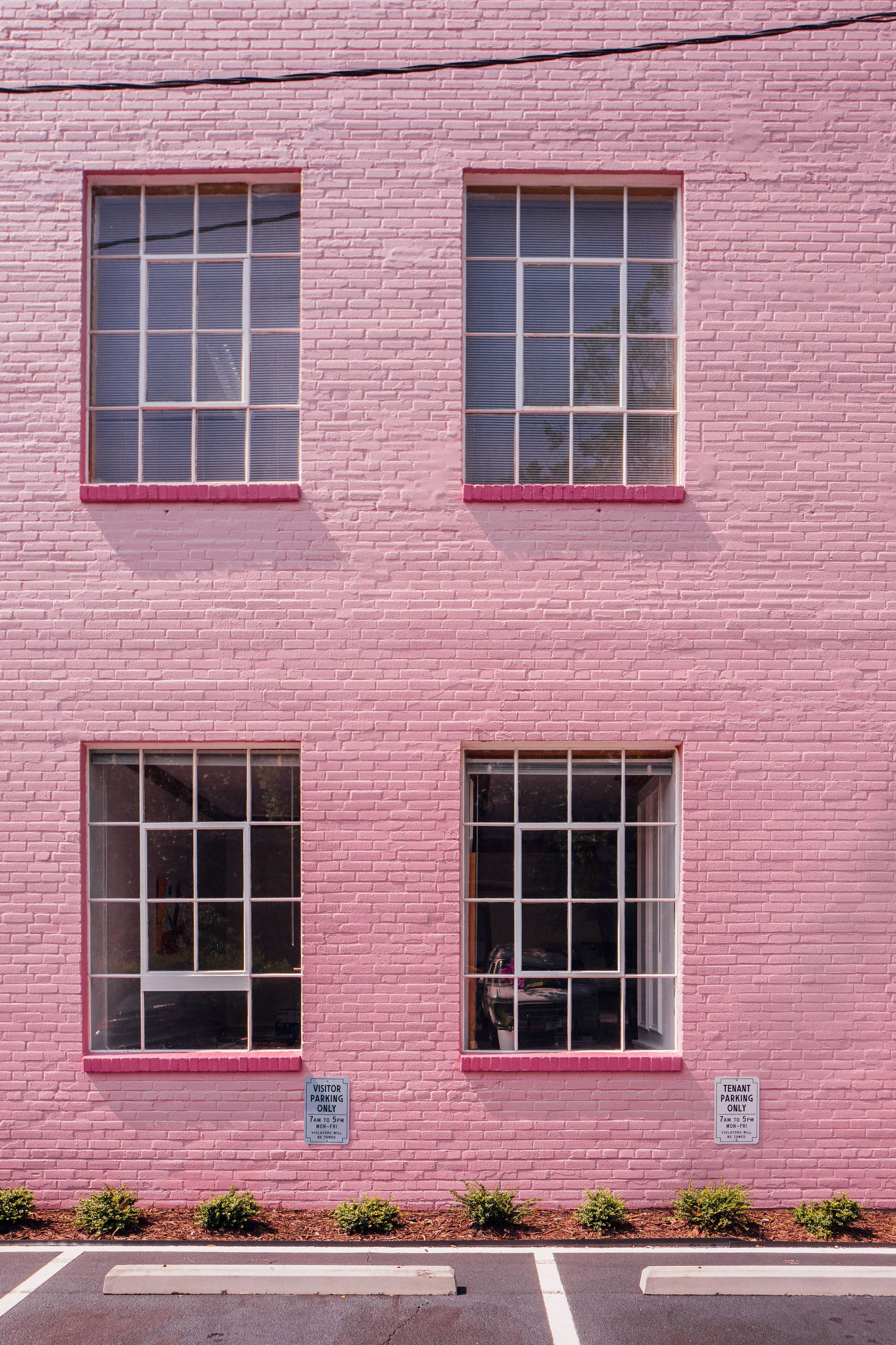 Pink brick building with four large windows, two on each floor. Small green bushes at the base, parking space in front, and 