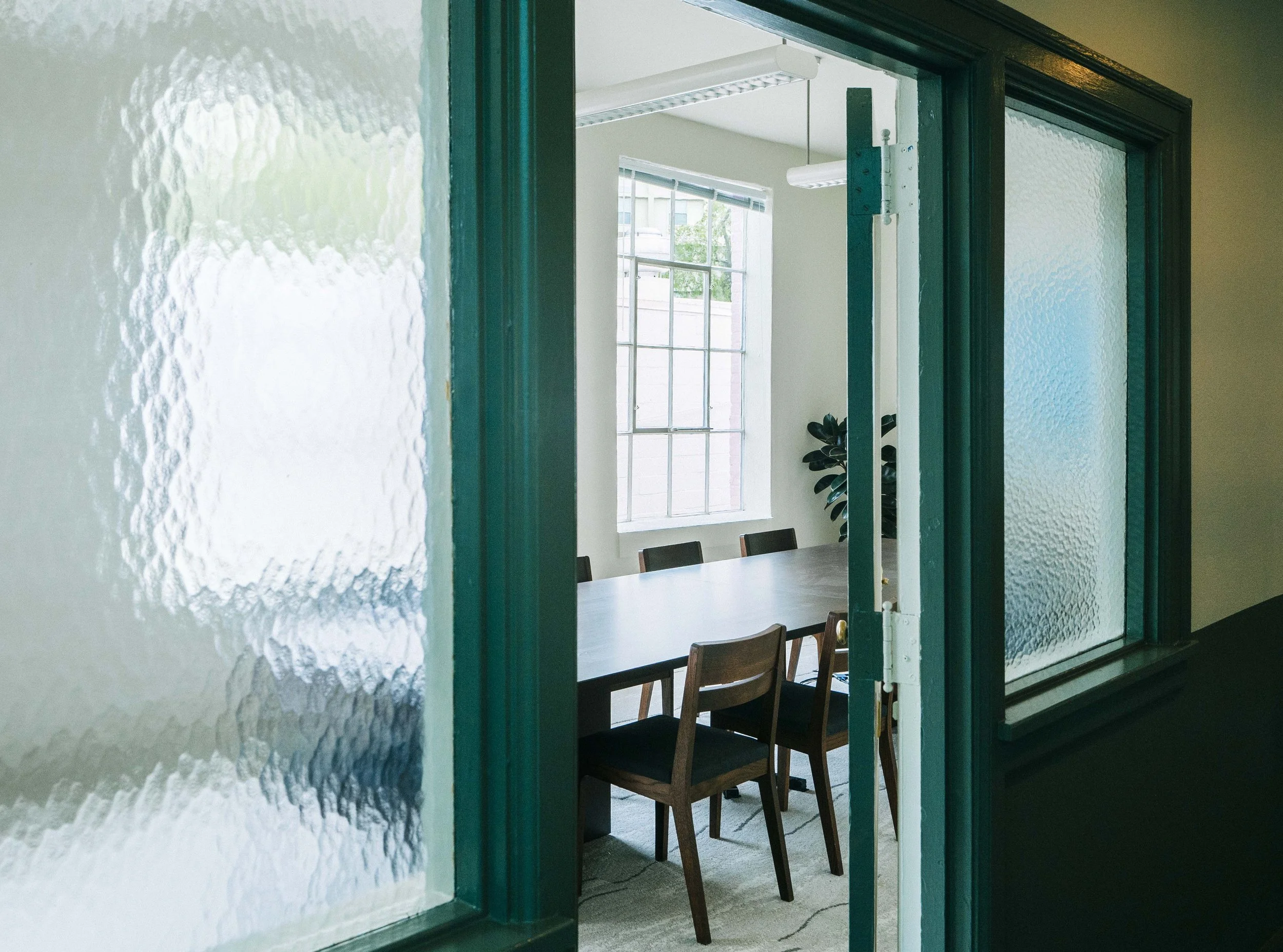 Interior view through a green-framed glass door into a bright conference room with large window, a wooden table, and chairs.