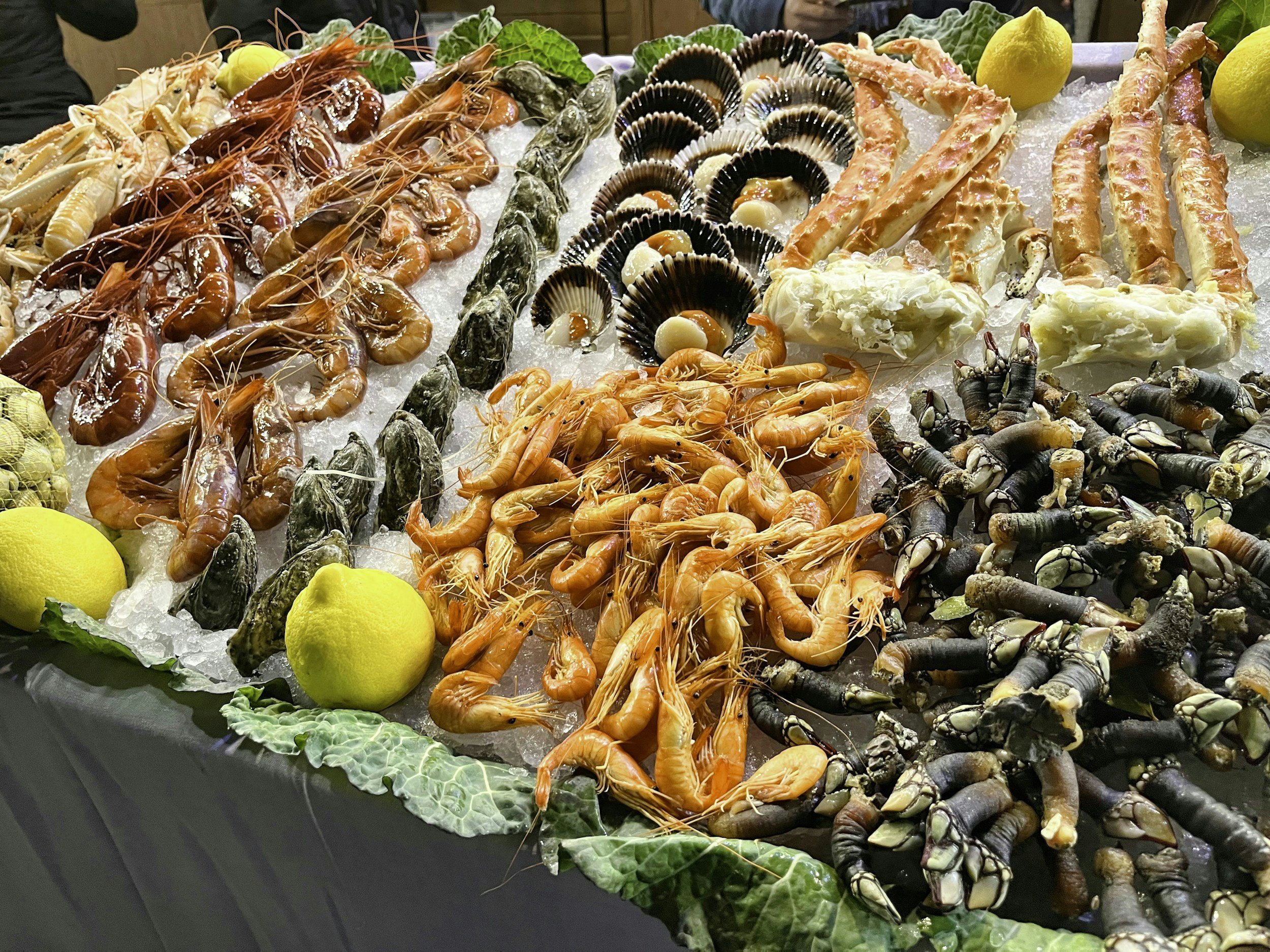 A seafood display with various fresh shellfish and crustaceans on ice, including shrimp, scallops, crab legs, and lobster claws, decorated with lemons and lettuce leaves.