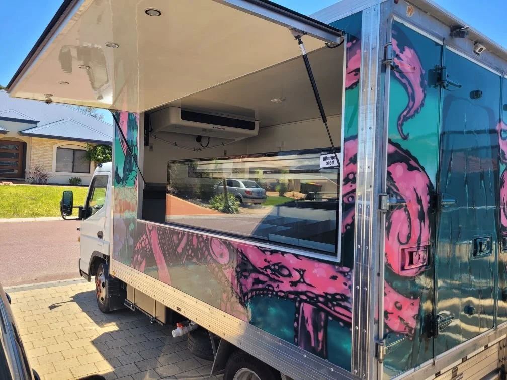 A colorful mobile food truck with pink and turquoise artwork, open serving window, parked on a street in a residential neighborhood.