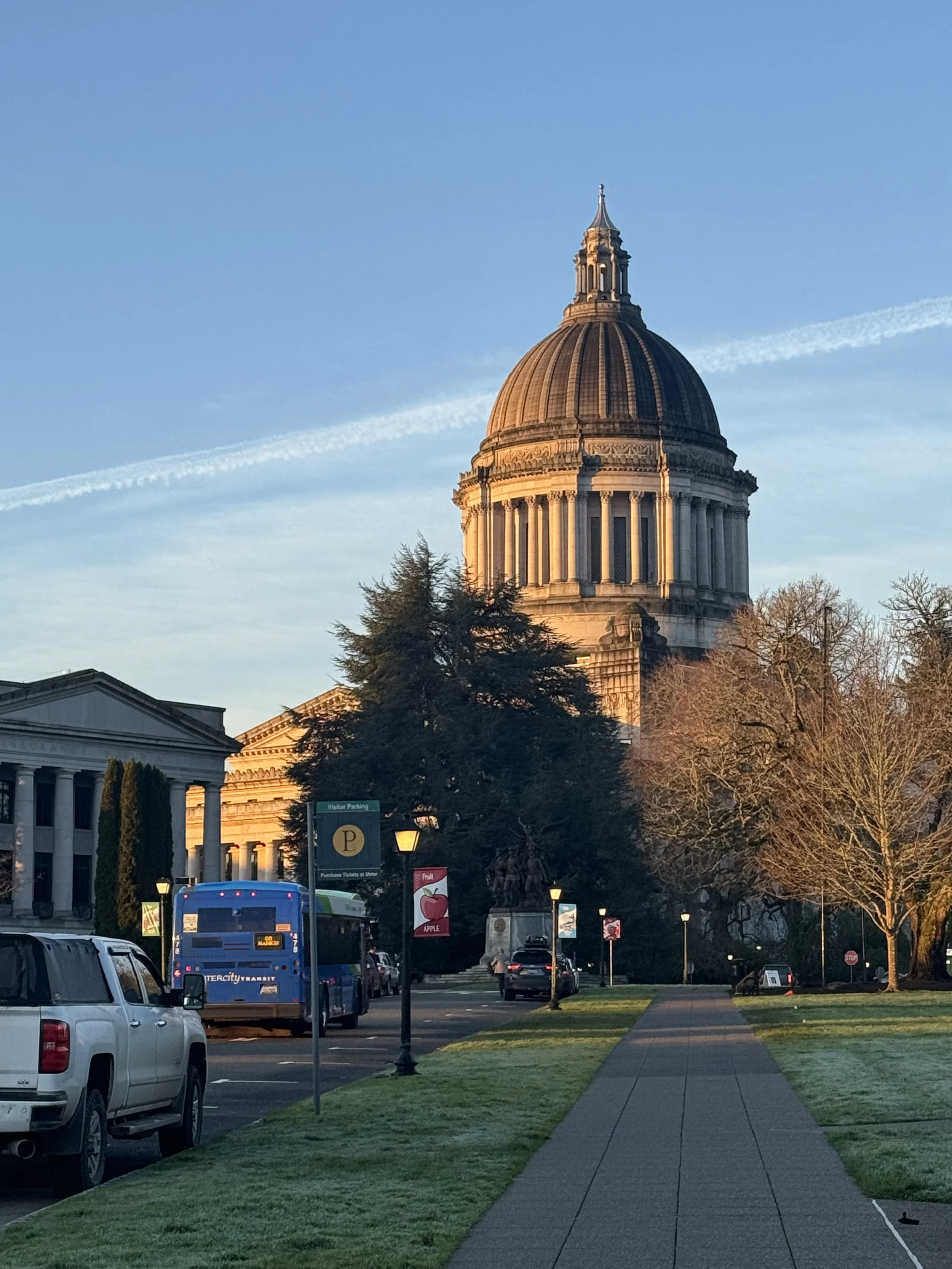 A city street with parked cars, a bus, and a sidewalk leading towards the large domed Washington State Legislative Building, with trees and clear blue sky in the background.