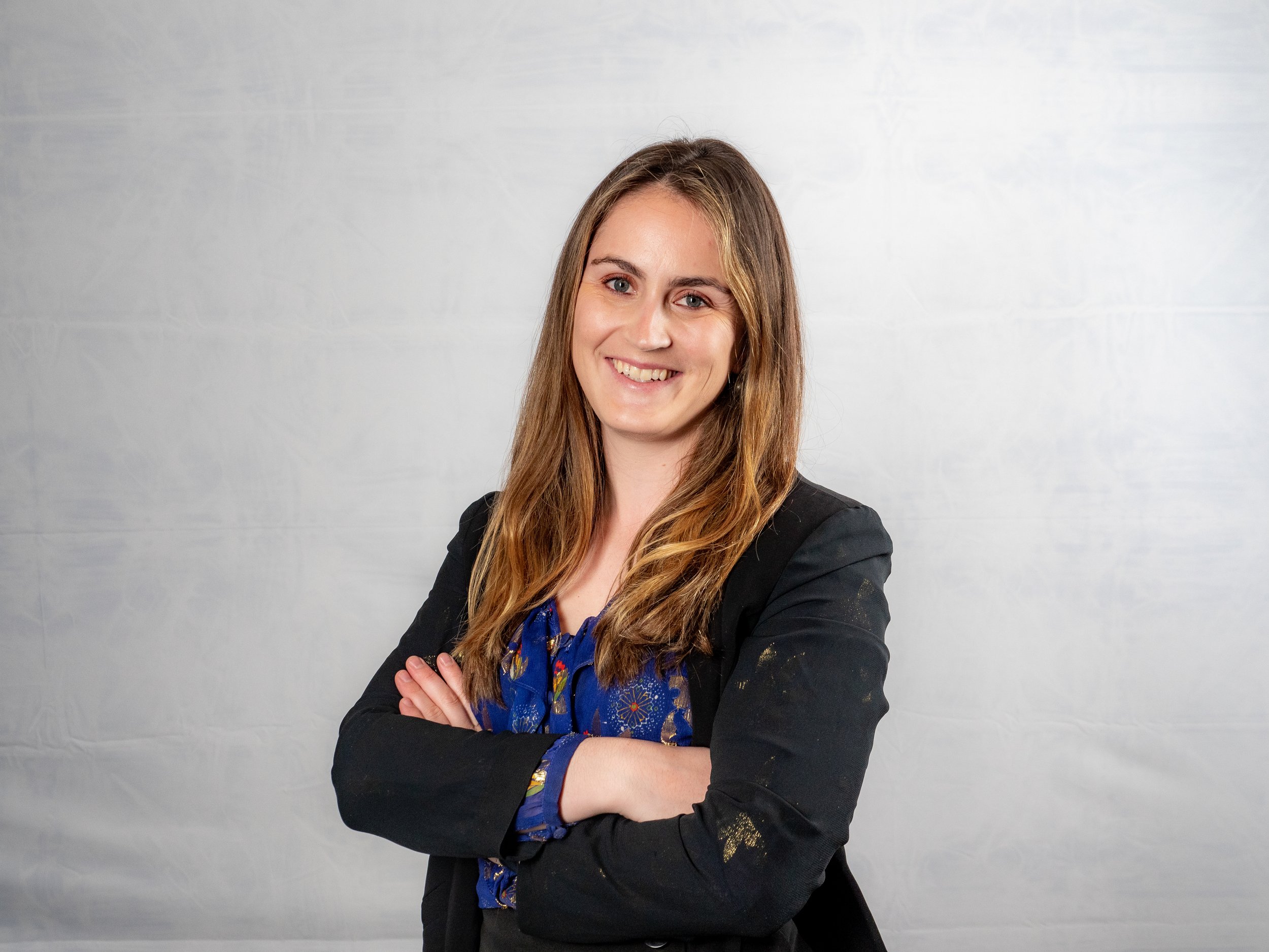 A smiling woman with long brown hair, crossed arms, wearing a black blazer over a blue blouse, standing against a light gray background.