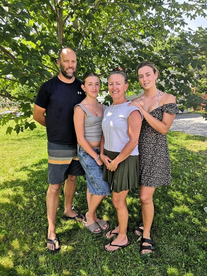 A family of four standing outdoors under a tree on a sunny day. The man is on the left, wearing a black t-shirt and shorts. The woman is in the middle, wearing a gray top and green skirt. The two young women are on the right, one wearing a gray tank top and denim skirt, the other in a black floral dress. All are smiling and posing for the photo.