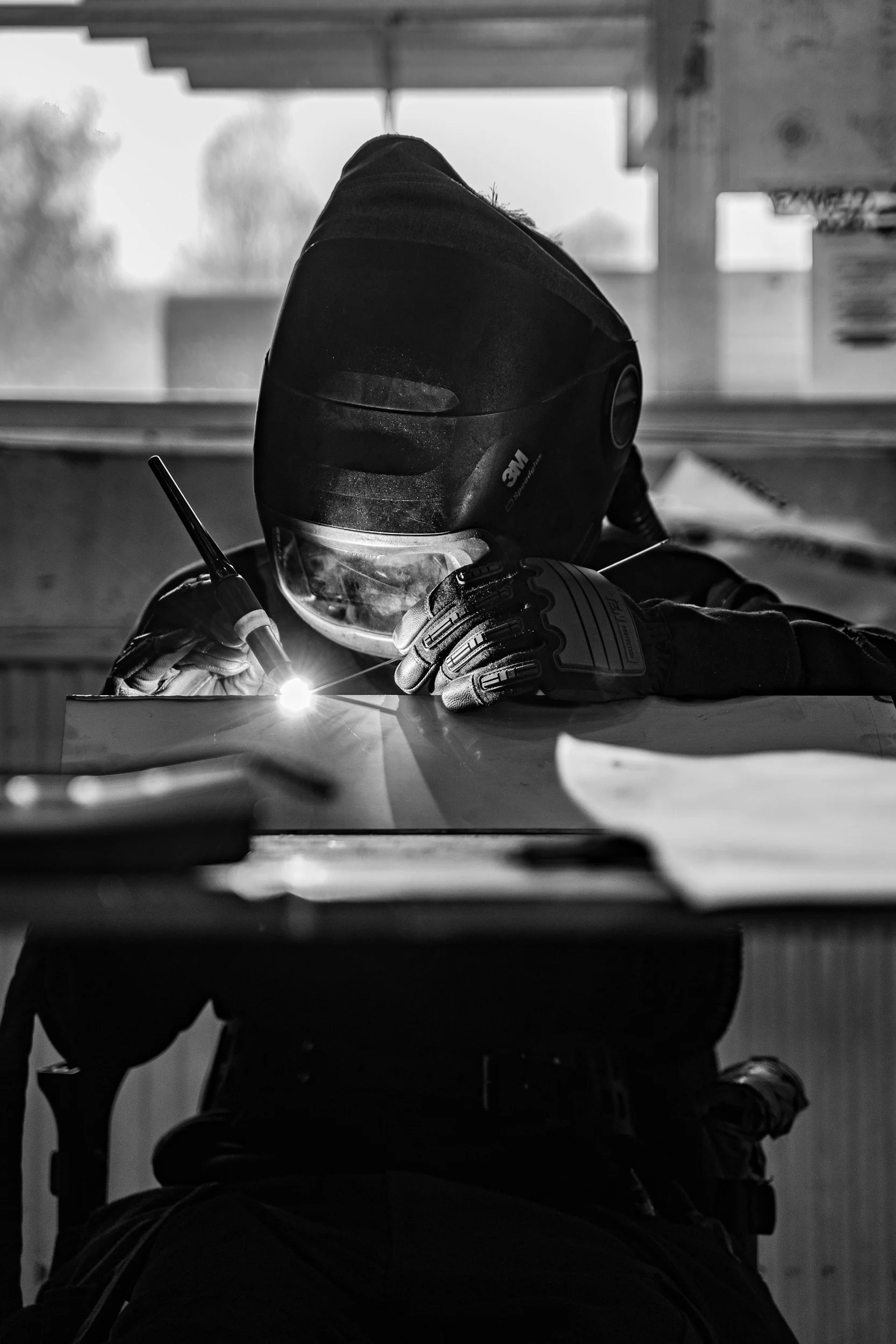 A person wearing a welding helmet and gloves is welding a piece of metal indoors, with sparks flying from the welding process.