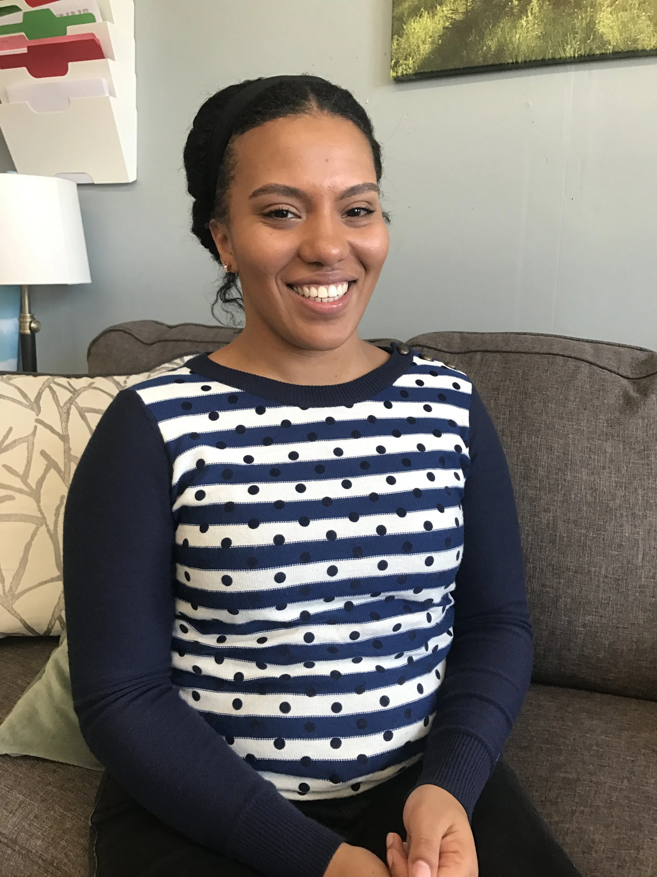 A woman sitting on a couch smiling, wearing a navy and white polka dot and striped long sleeve shirt, with a beige pillow and a dark sofa behind her, and a landscape picture hanging on the wall.