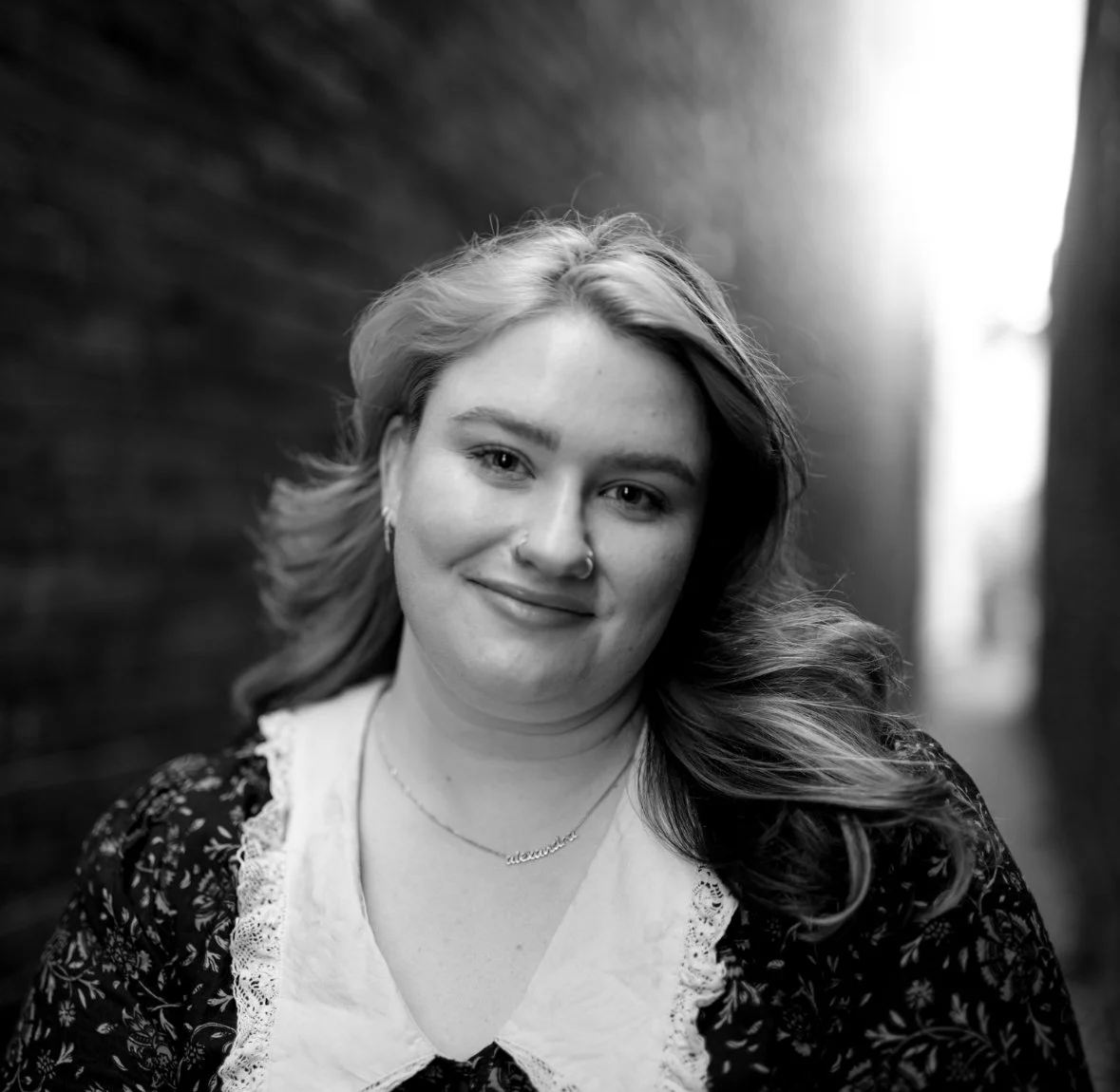 Black and white portrait of a smiling young woman with wavy hair, wearing a patterned top, a necklace, and earrings, standing between two brick walls with light shining in the background.