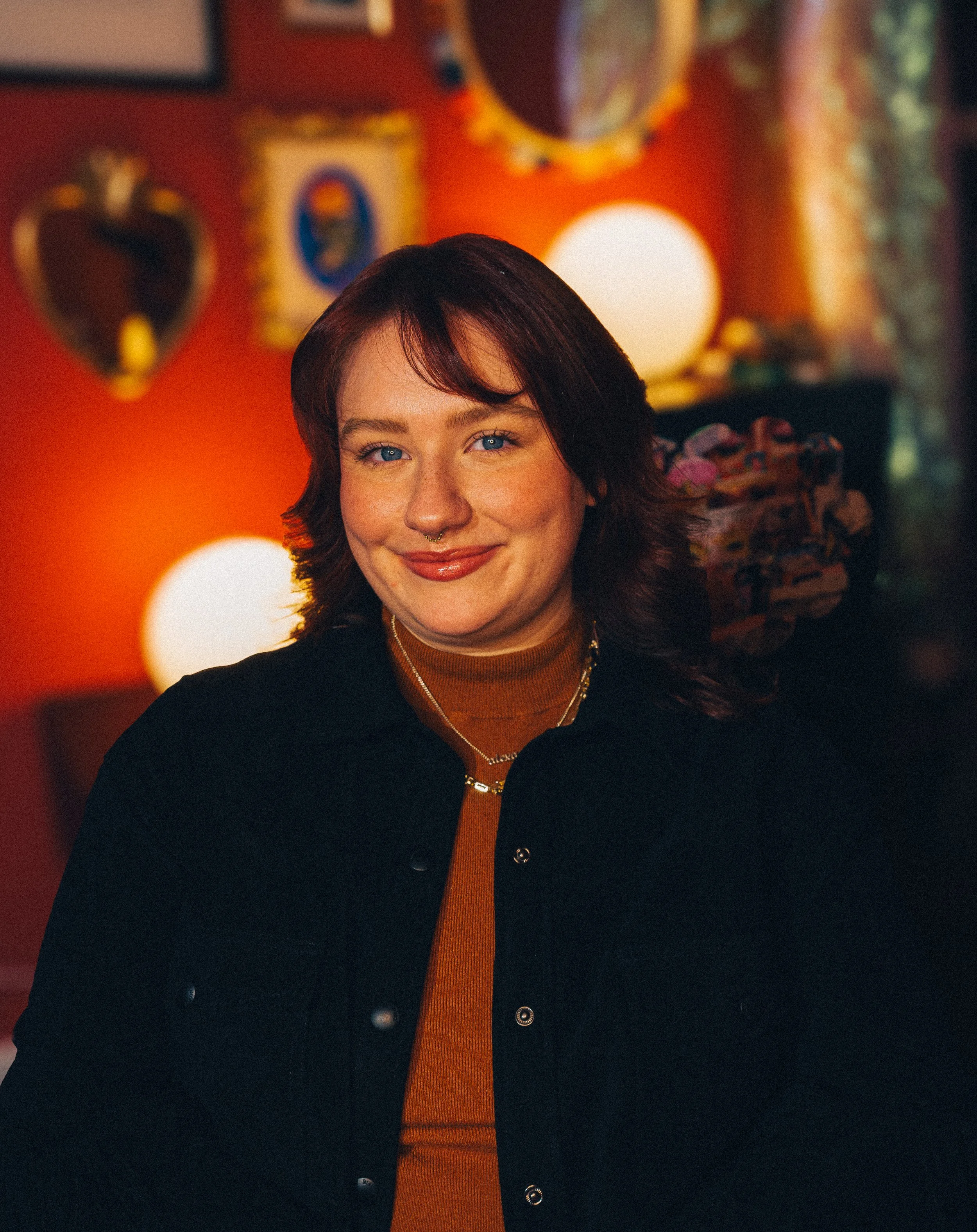 A woman with shoulder-length dark hair, freckles, a septum piercing, and blue eyes smiles at the camera. She is wearing a black jacket over a brown turtleneck and layered necklaces. The background features warm lighting, heart-shaped mirrors, and framed pictures on a red wall.