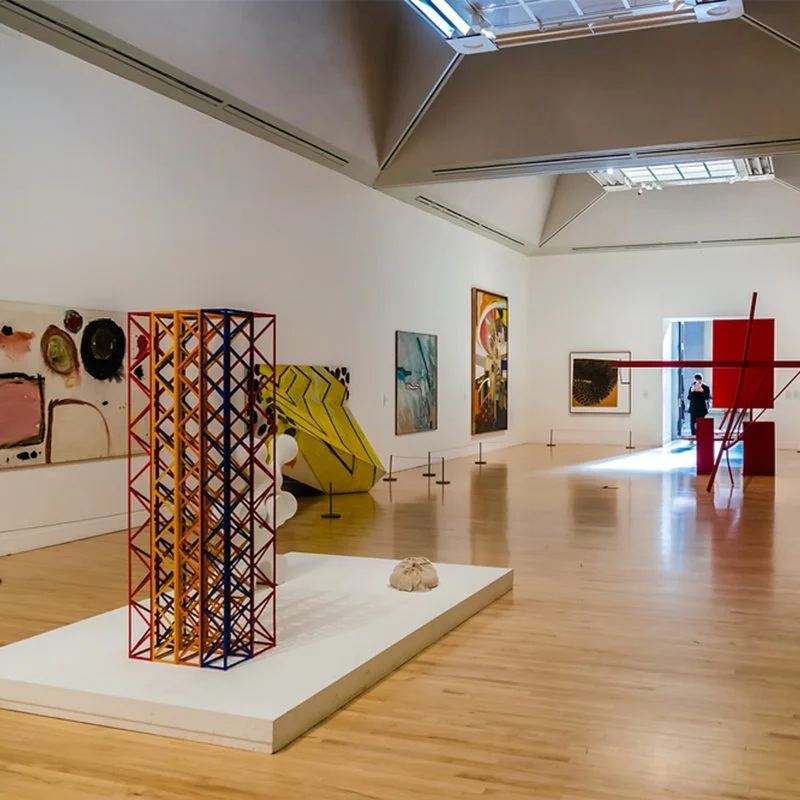 Art gallery exhibition with various sculptures and paintings on white walls, featuring abstract and modern art pieces, with some visitors viewing the artwork.