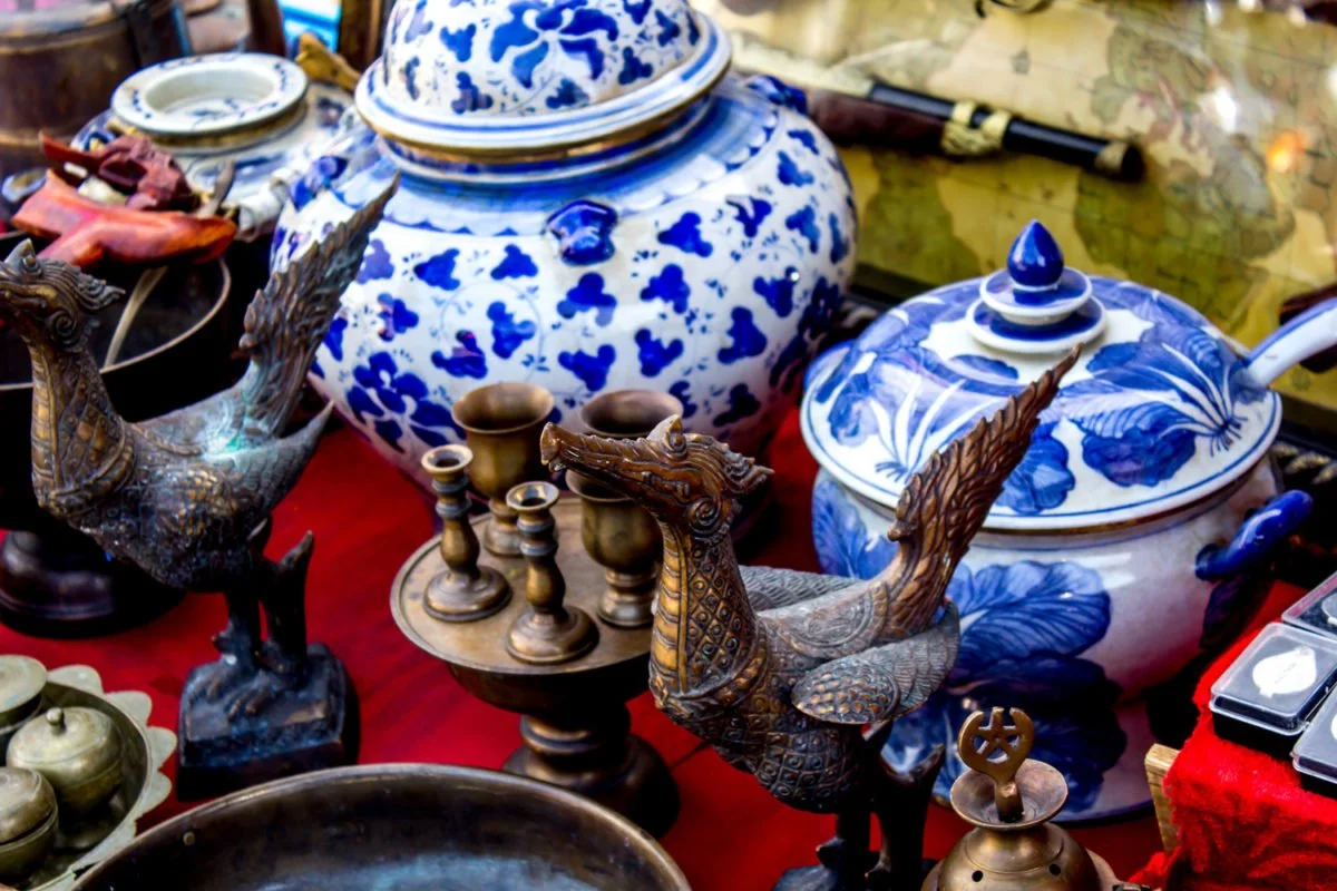 A display of antique Asian-style decorative objects including large blue and white porcelain jars, ornate brass sculptures of phoenix-like birds, small brass vases, and intricate brass figurines, all arranged on a red tablecloth.