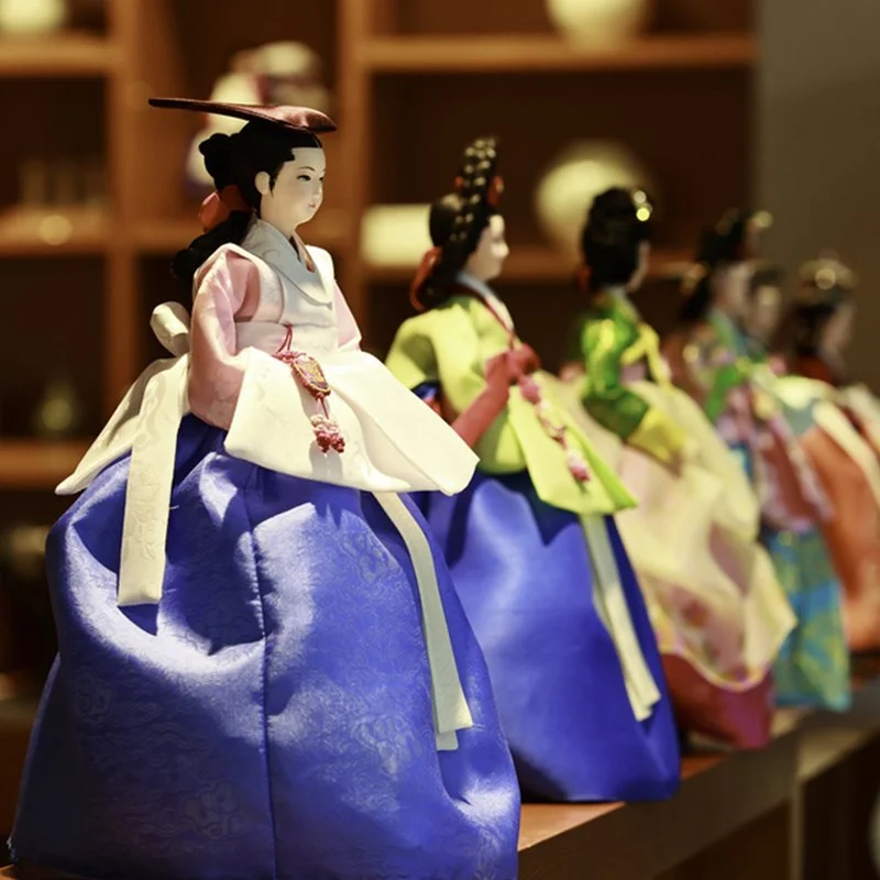 A row of traditional Korean dolls dressed in colorful hanbok, displayed on a shelf with a blurred background.
