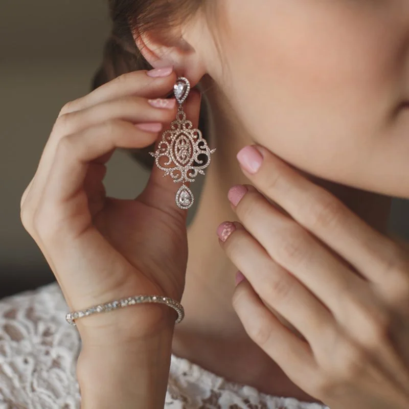 Close-up of a woman holding an ornate diamond earring near her ear, wearing a matching diamond bracelet on her wrist, with painted nails and lace clothing visible
