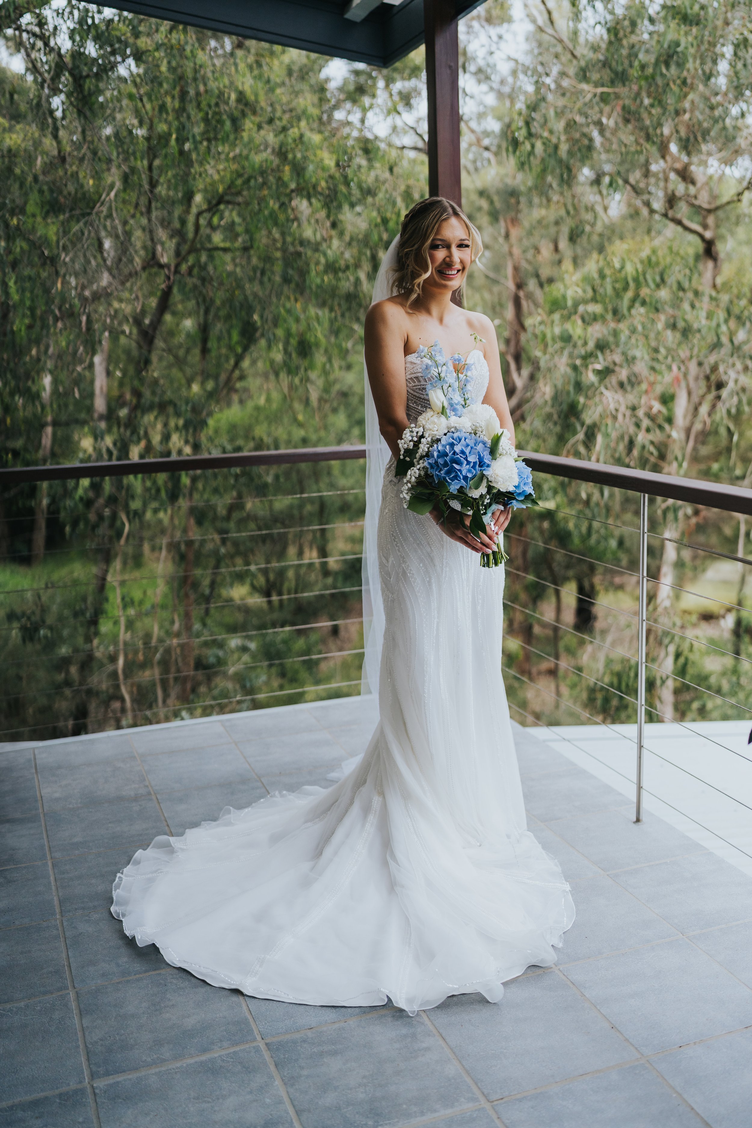 Bride in a white wedding dress holding a blue and white bouquet, standing on a balcony with a forest background.