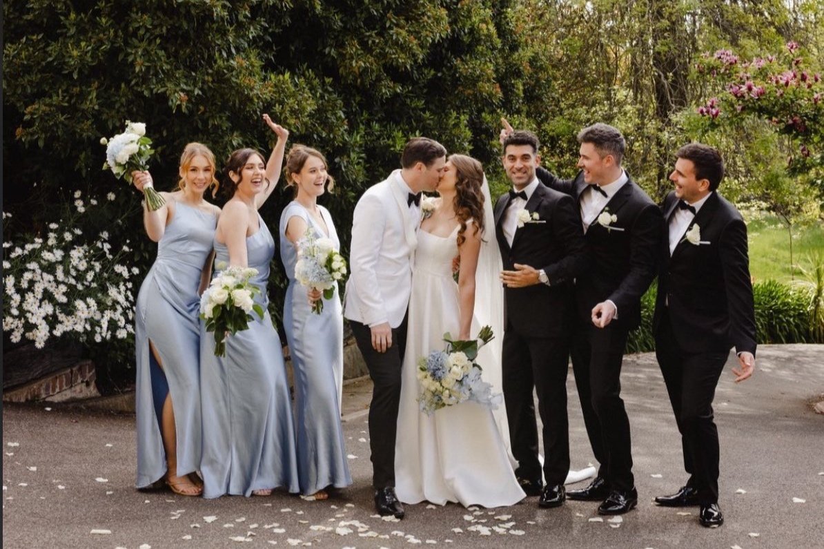 A wedding party with the bride and groom kissing, surrounded by bridesmaids in light blue dresses and groomsmen in black tuxedos, outdoors in a garden setting.