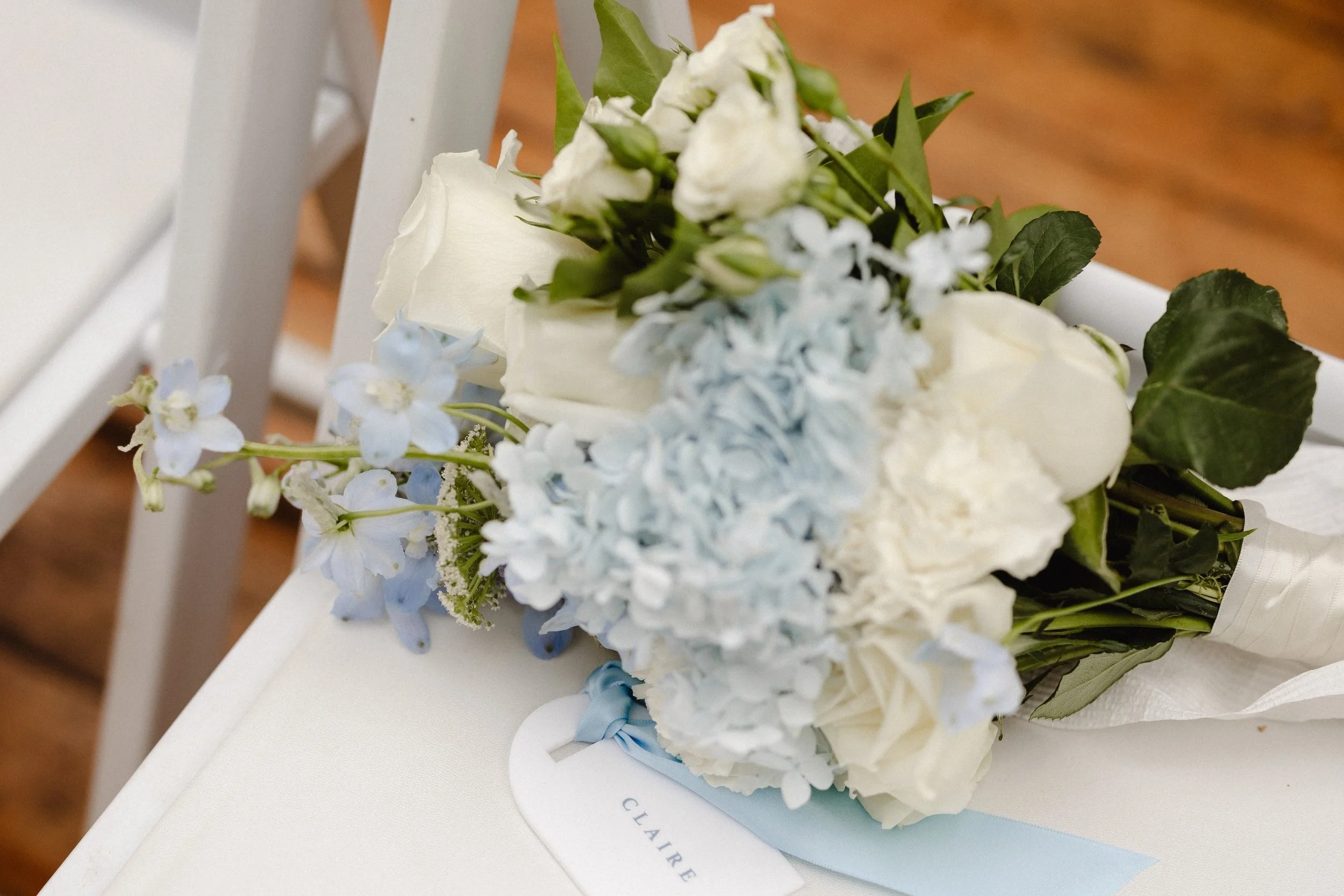 A bouquet of white and light blue flowers placed on a white chair with a blue ribbon and a tag reading 'Claire'