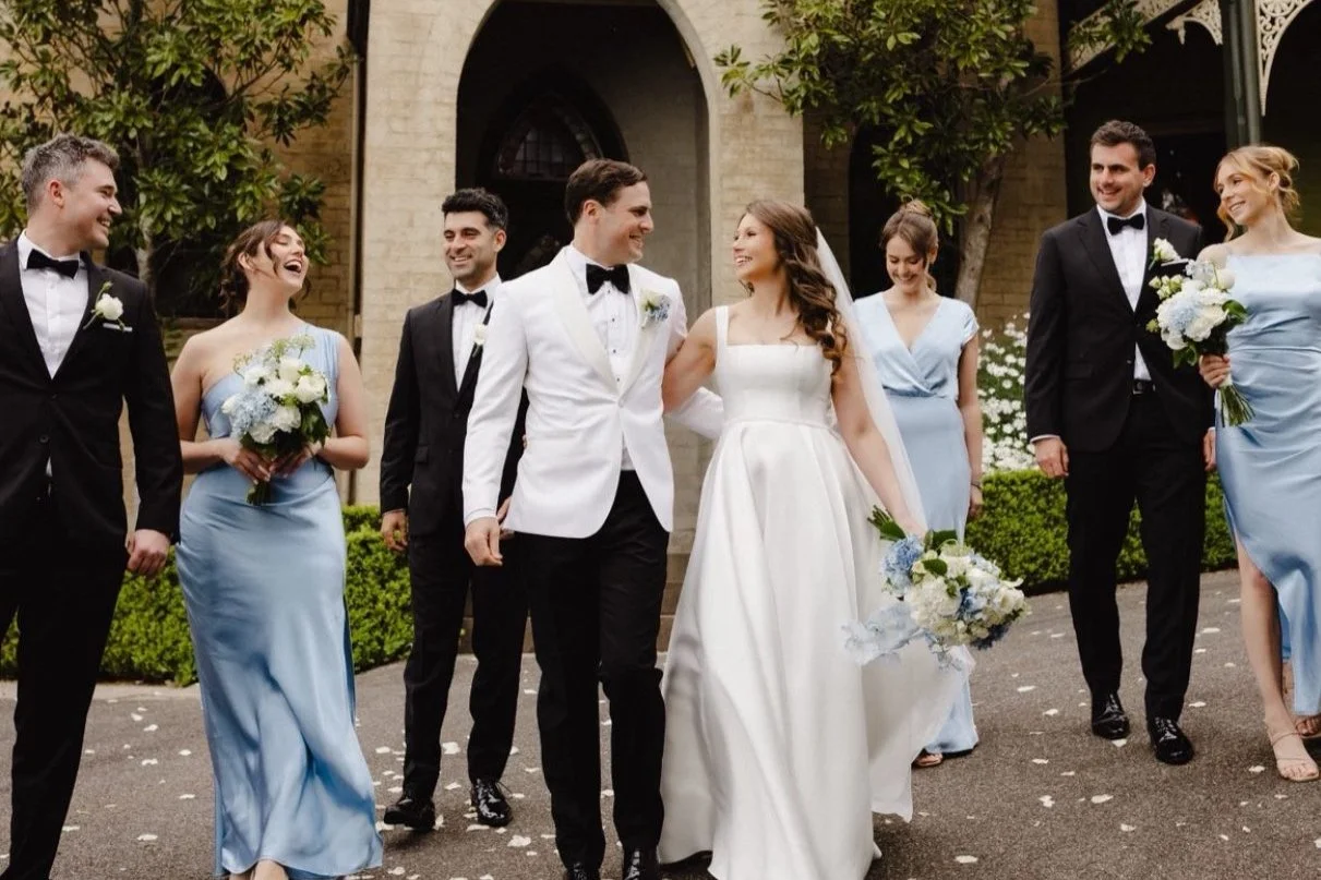 A wedding party walking outside in formal attire, with the bride and groom in the center, surrounded by bridesmaids and groomsmen, holding bouquets and smiling.