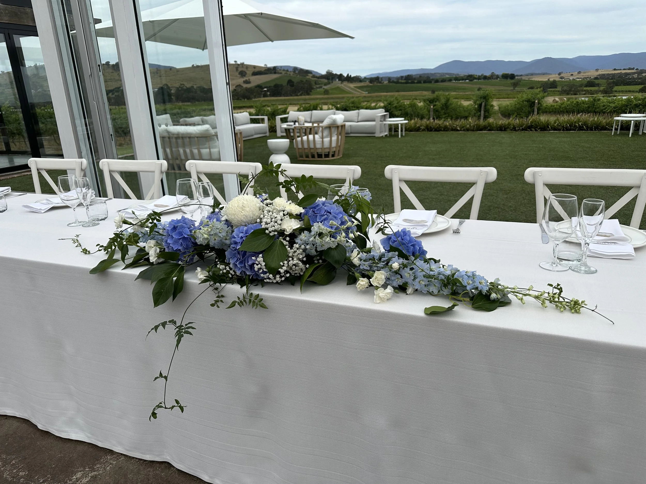 A long white table decorated with a central floral arrangement of blue hydrangeas, white roses, and greenery, set for a formal event with glasses, plates, and napkins. In the background, an outdoor patio with white furniture and a scenic view of rolling green hills and mountains under a cloudy sky.