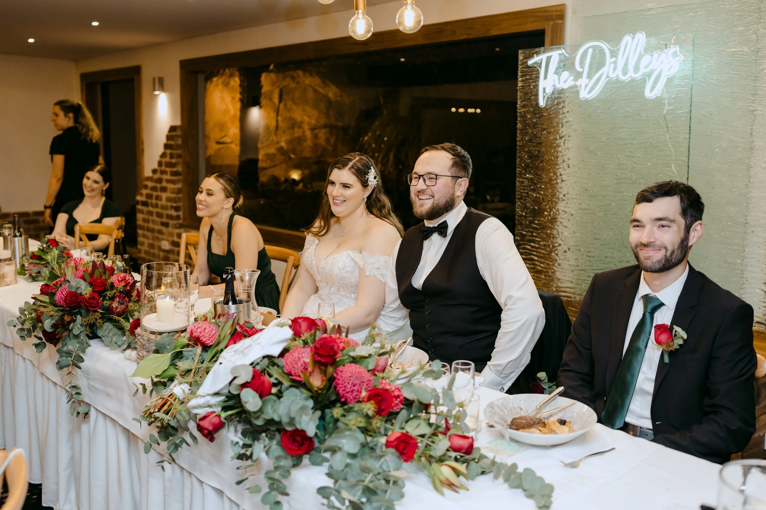 Wedding reception table with six people, including a bride in a white dress and three men in suits, seated behind a floral centerpiece with red and pink flowers. There are wine bottles, glasses, and plates on the table. A neon sign on the wall reads 'The Dulles'.