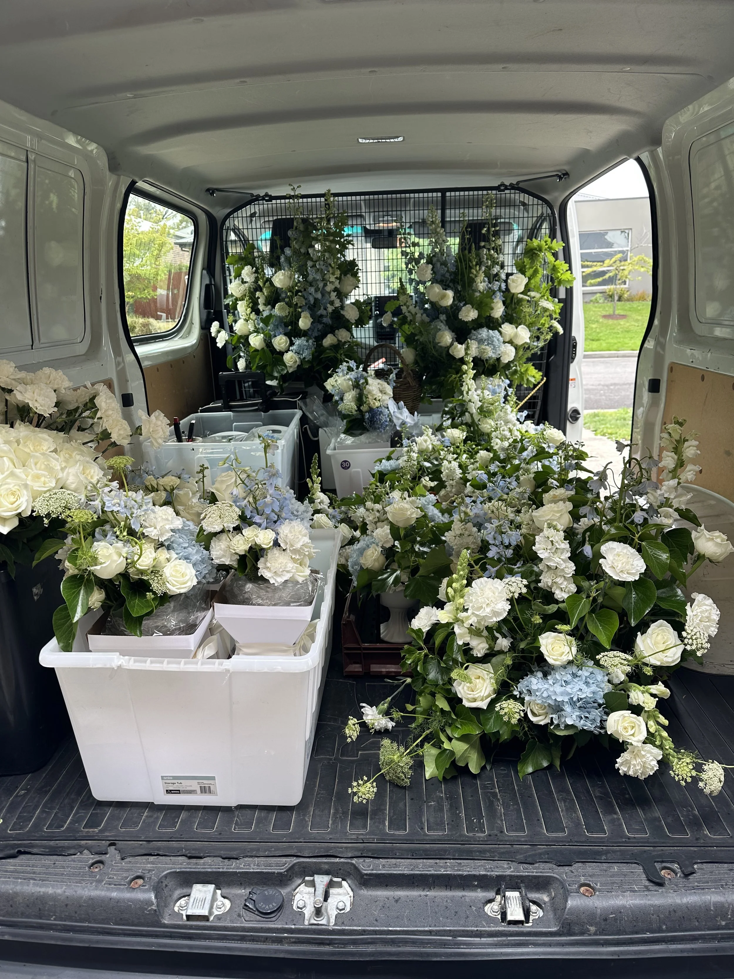 Inside a vehicle loaded with white and blue flower arrangements for a funeral or event.